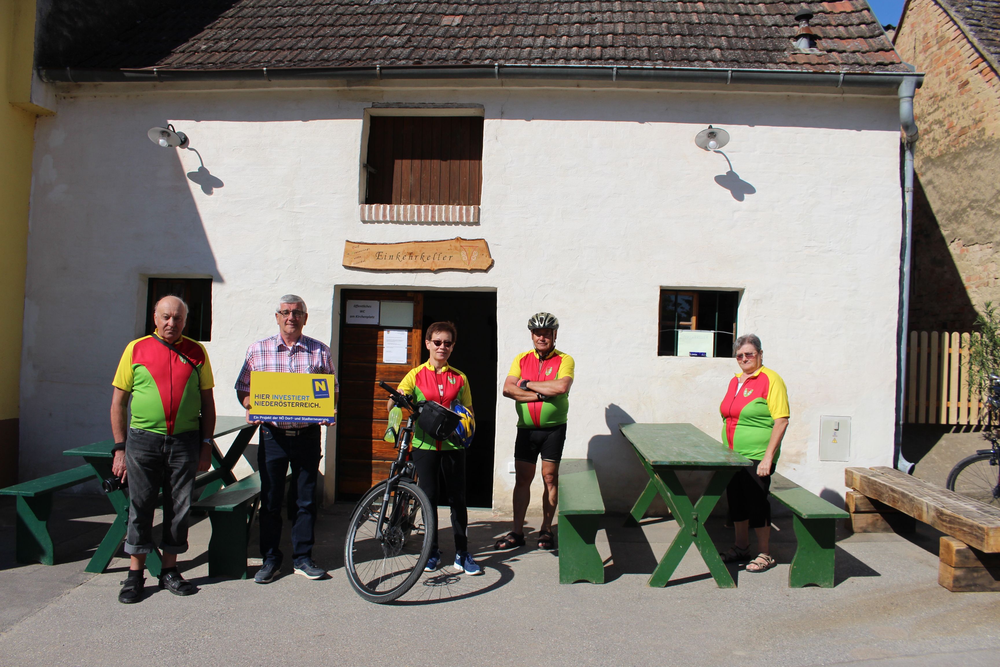 Před restaurací „Einkehrkeller Ottenthal“ stojí skupina pěti lidí. Dva z nich mají na sobě cyklistické dresy, jeden drží kolo. Tabule upozorňuje na investice v Dolním Rakousku.