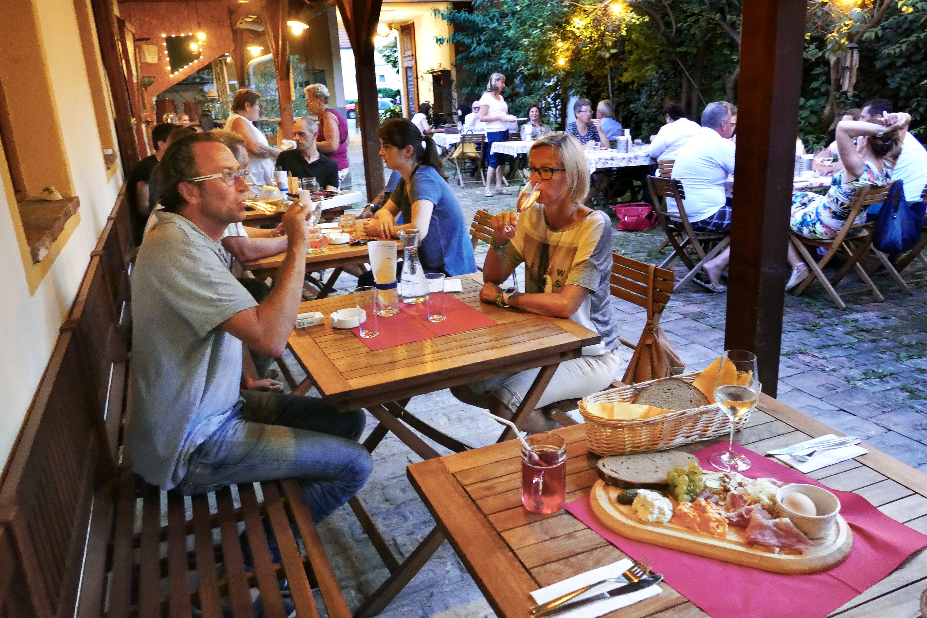 Lidé sedí u dřevěných stolů v útulném venkovním prostoru a vychutnávají si jídlo a nápoje.