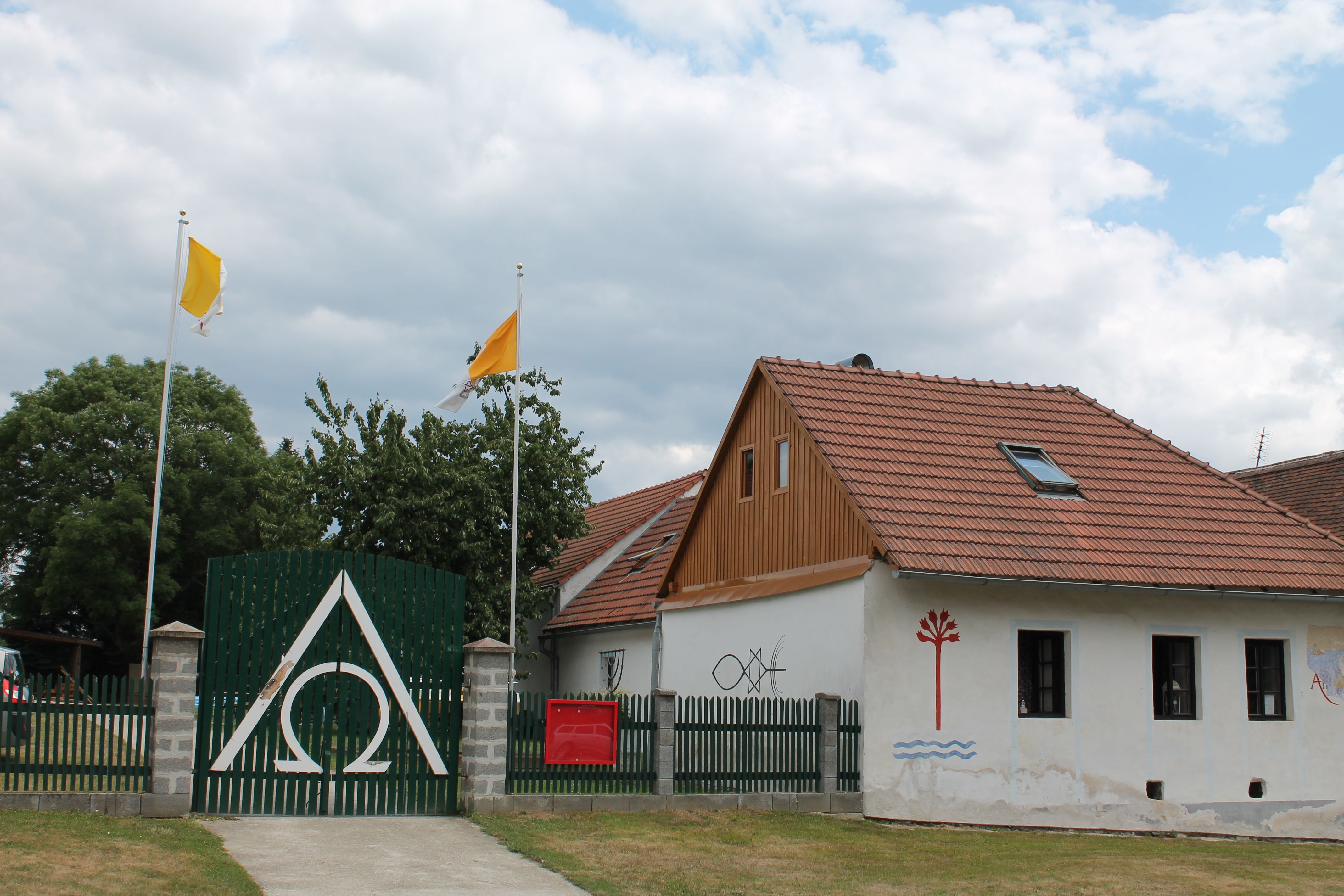 Dům s červenými střešními taškami, zelený plot se symbolem Alfa Omega, dvě žluté vlajky a nástěnné malby.