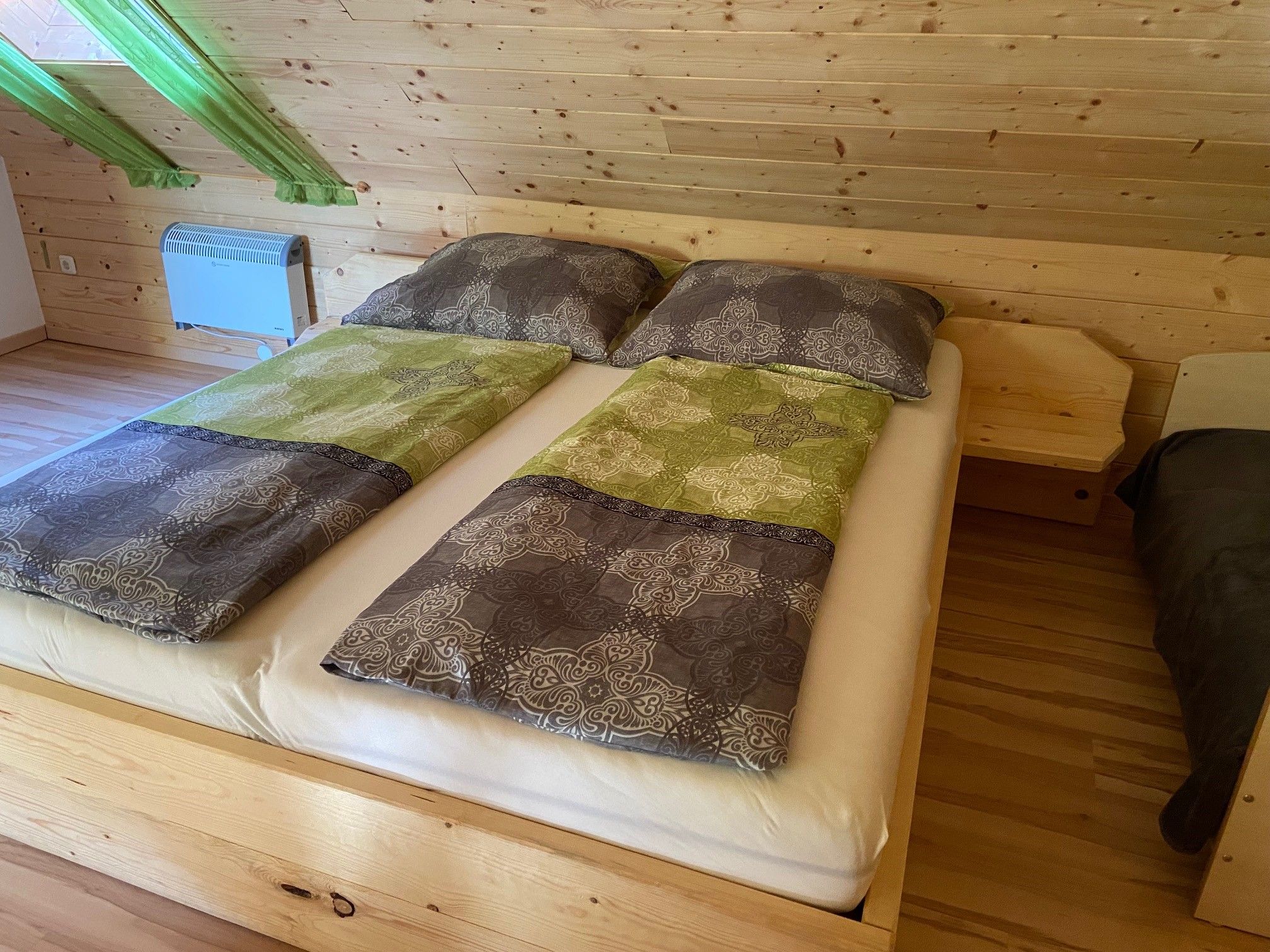 Manželská postel v pokoji s dřevěným obložením a zelenými závěsy.