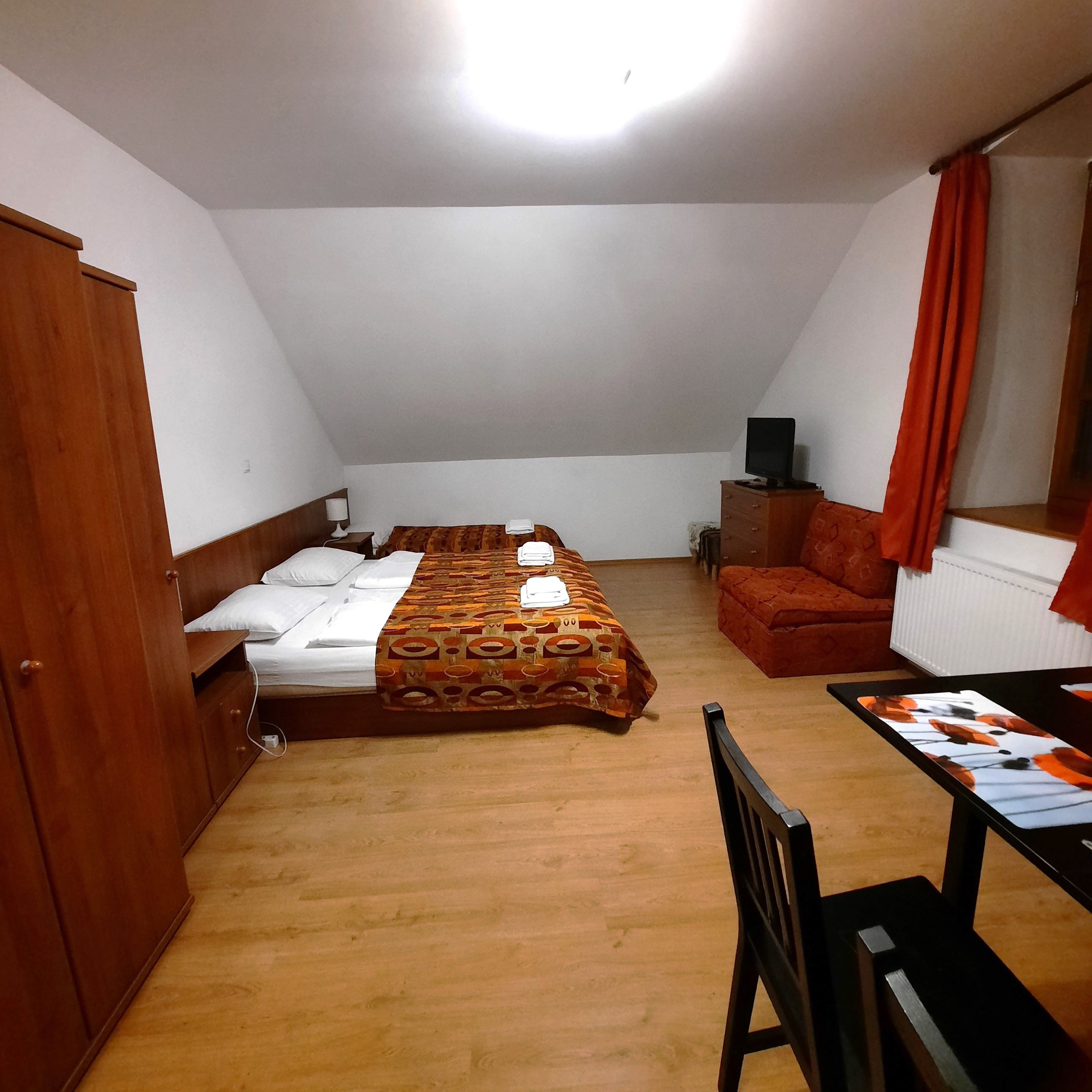 Jednoduše zařízený pokoj s manželskou postelí, pohovkou, stolem a židlí. Pokoji dominuje dřevěný nábytek a oranžové závěsy.