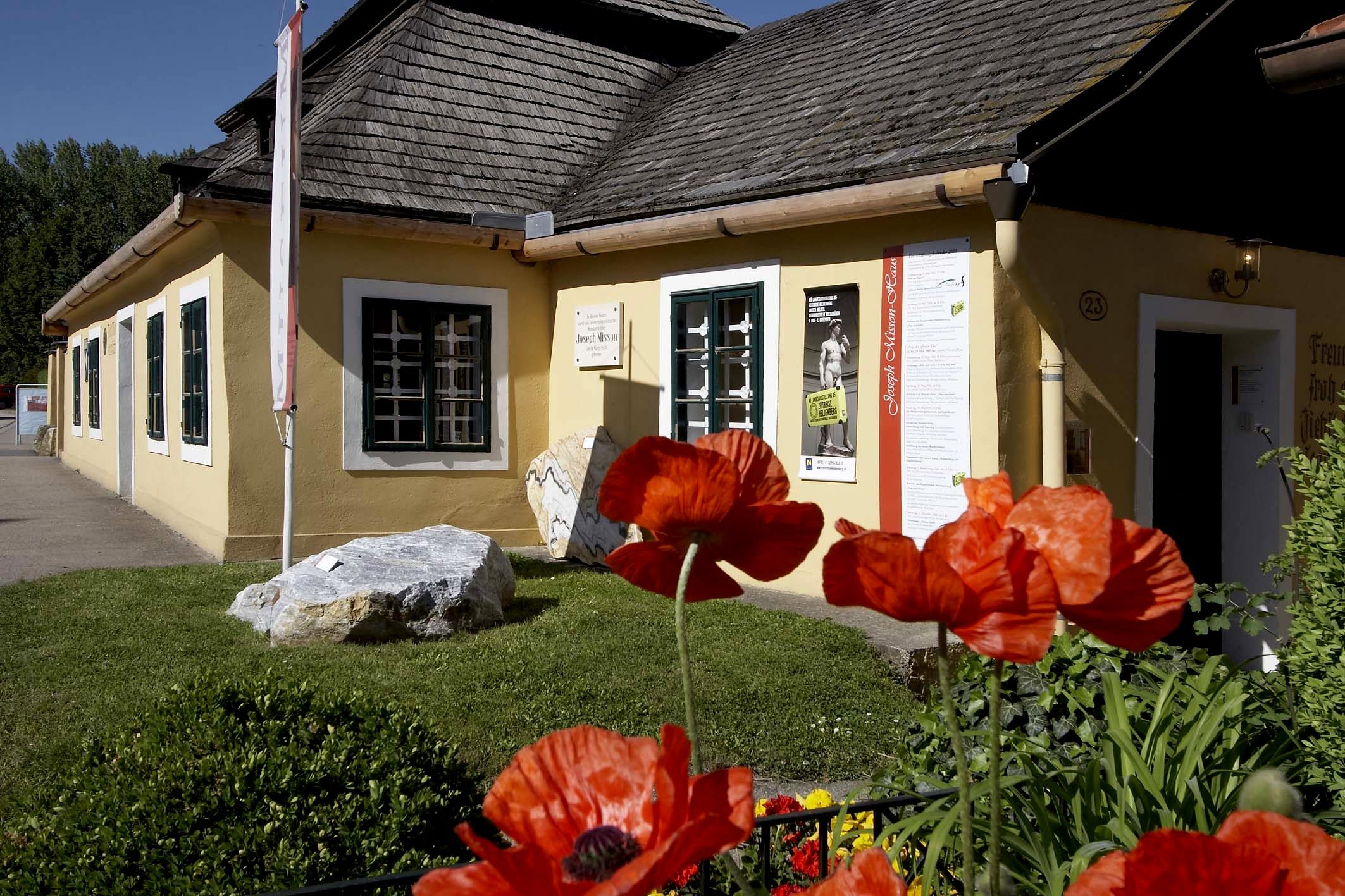 Žlutý dům se šindelovou střechou a máky v popředí.