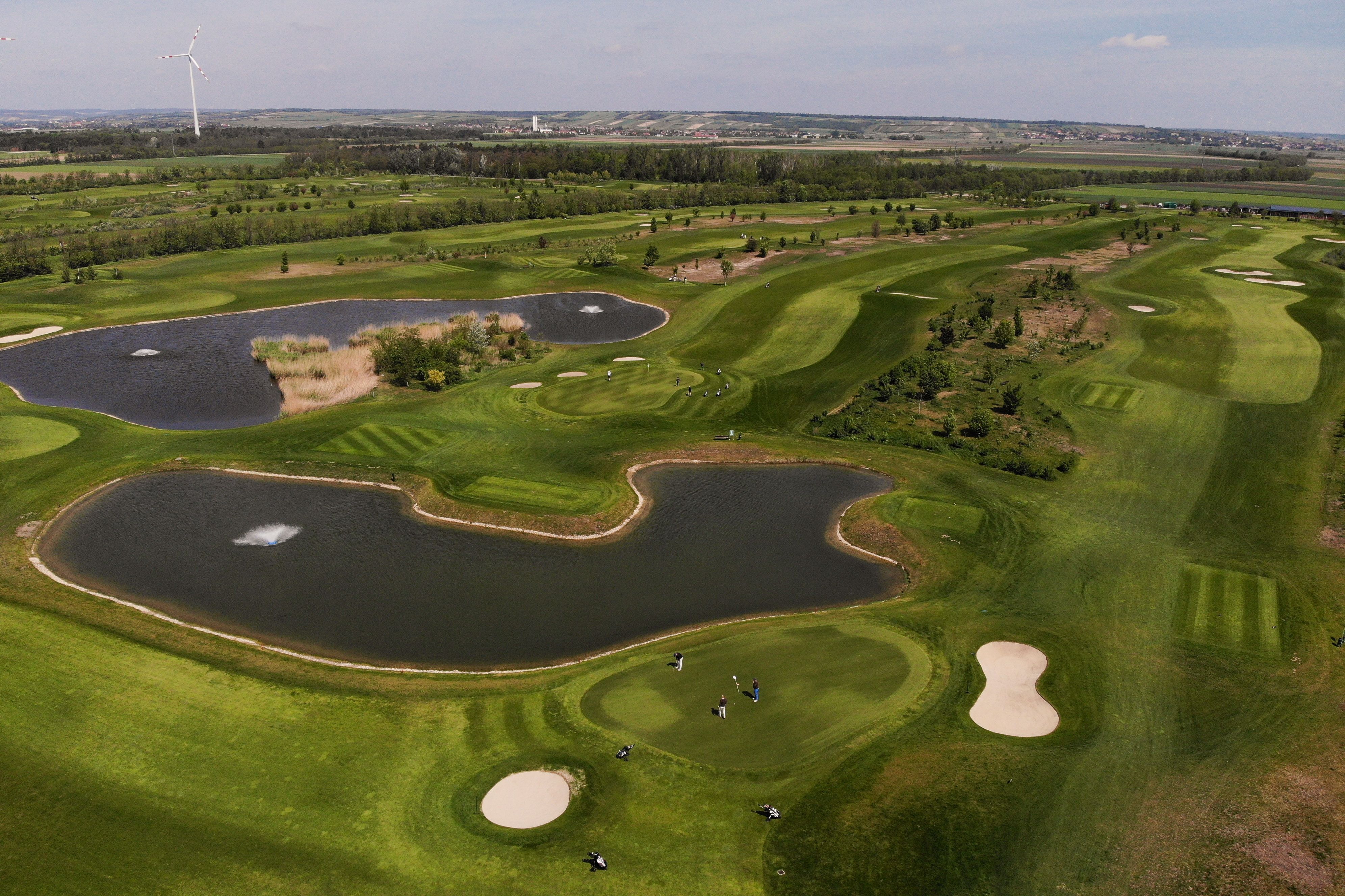Letecký pohled na golfové hřiště s rybníky a větrnou turbínou v pozadí.