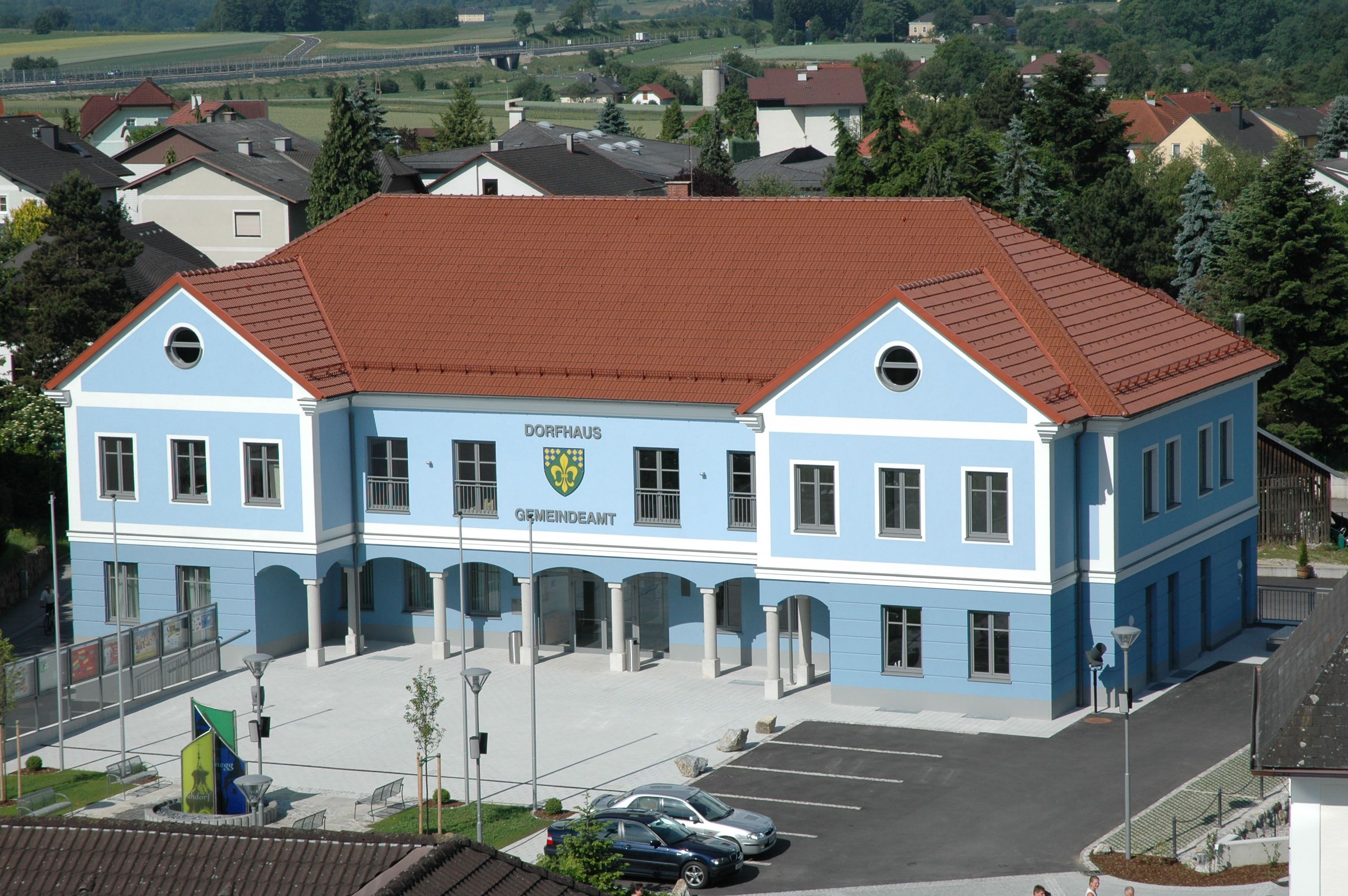 Modrá budova s červenou střechou, označená jako obecní úřad, obklopená domy a stromy.
