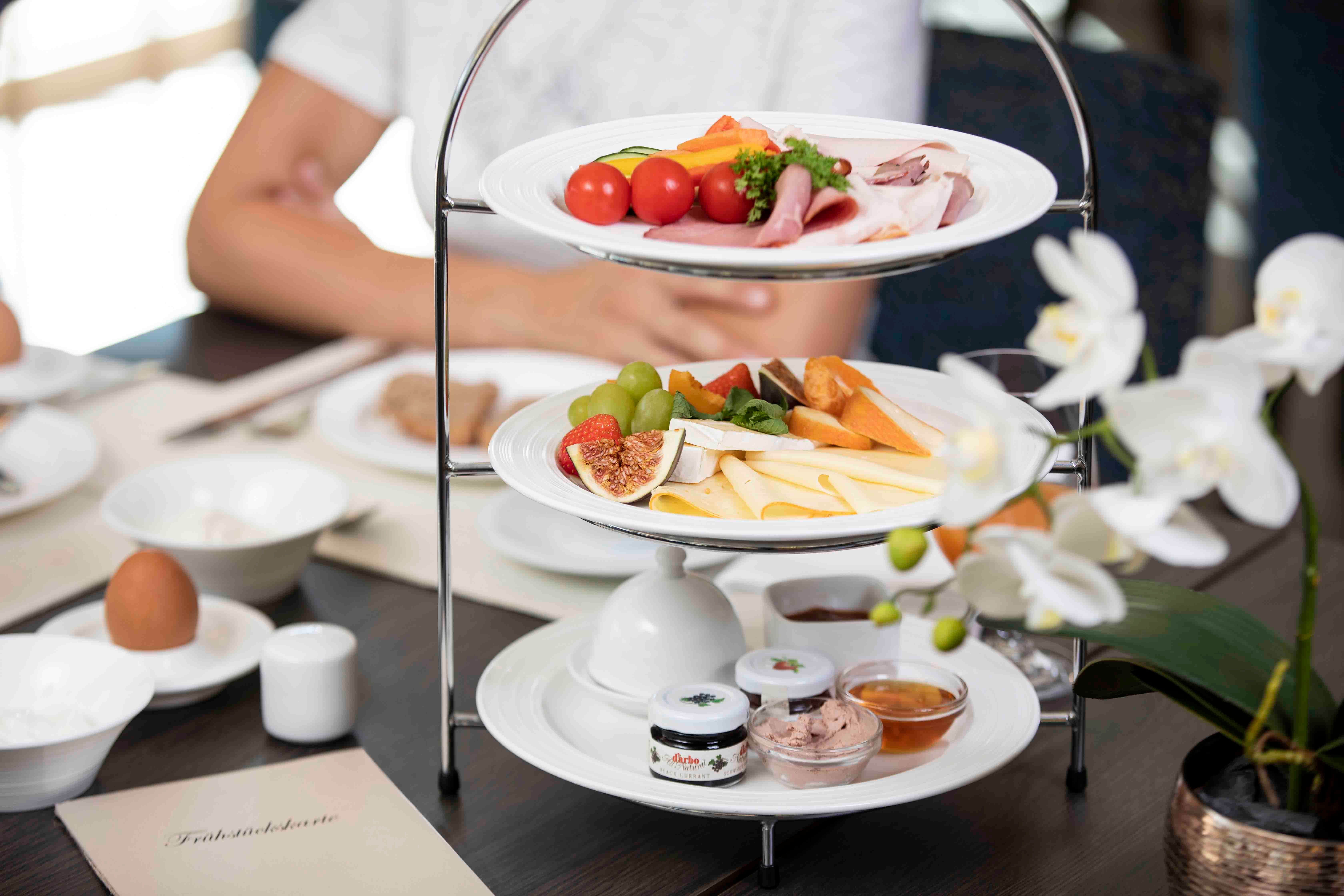 Snídaňový stůl s etáží plnou sýrů, uzenin, ovoce a pomazánek. V popředí bílá orchidej.