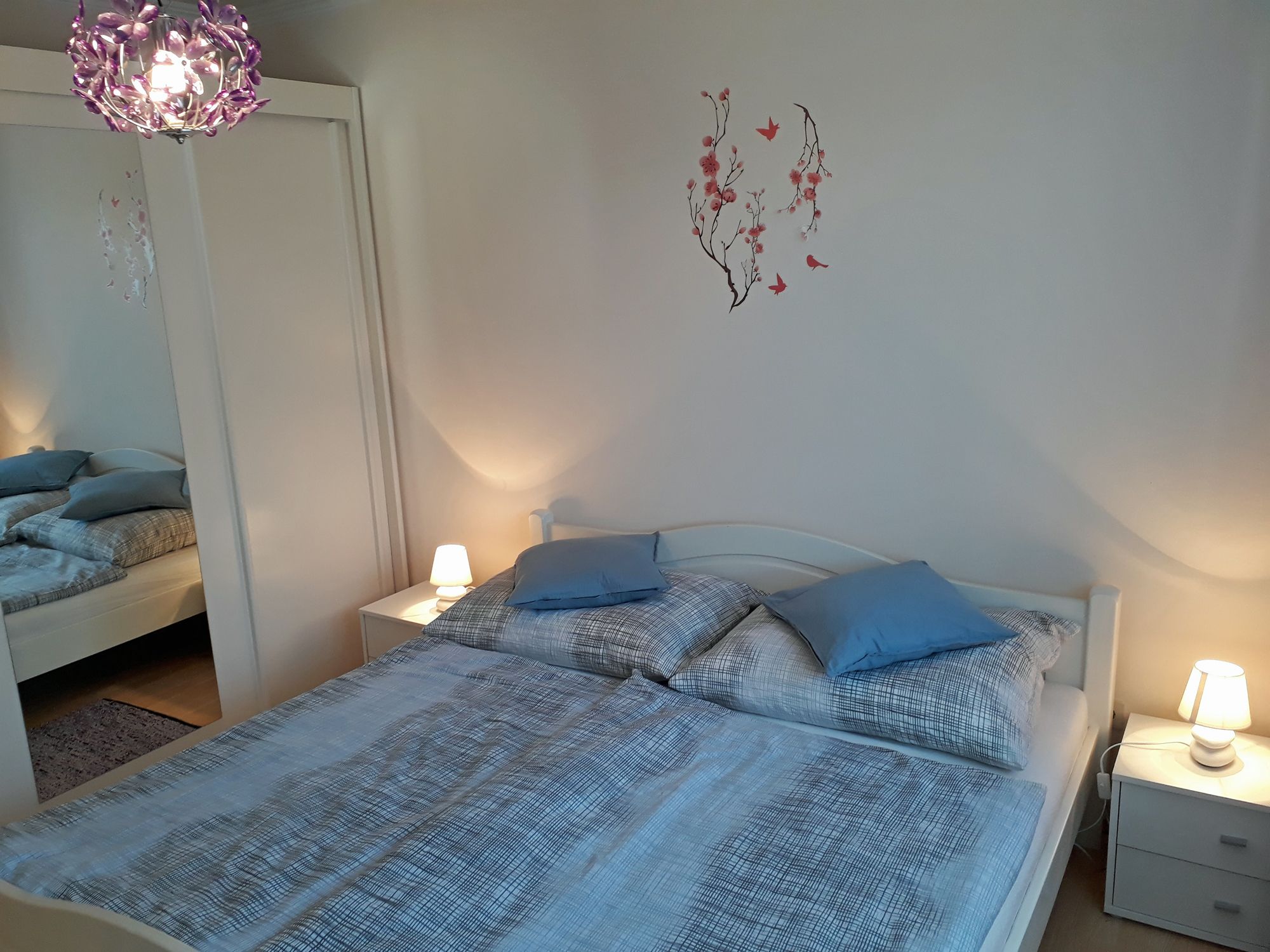 Útulná ložnice s manželskou postelí, modrým ložním prádlem, nočními stolky s lampičkami a nástěnným tetováním s květinami a ptáky.