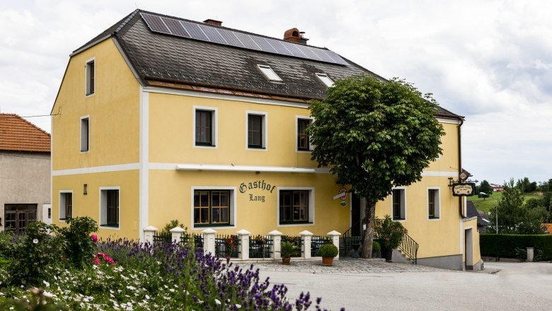 Žlutý penzion se solárními panely na střeše, obklopený květinami a stromy.