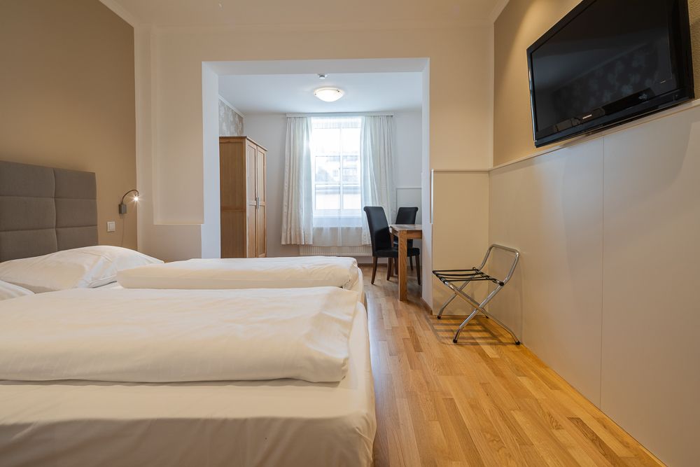 Hotelový pokoj se dvěma lůžky, televizí, stolem a židlemi.