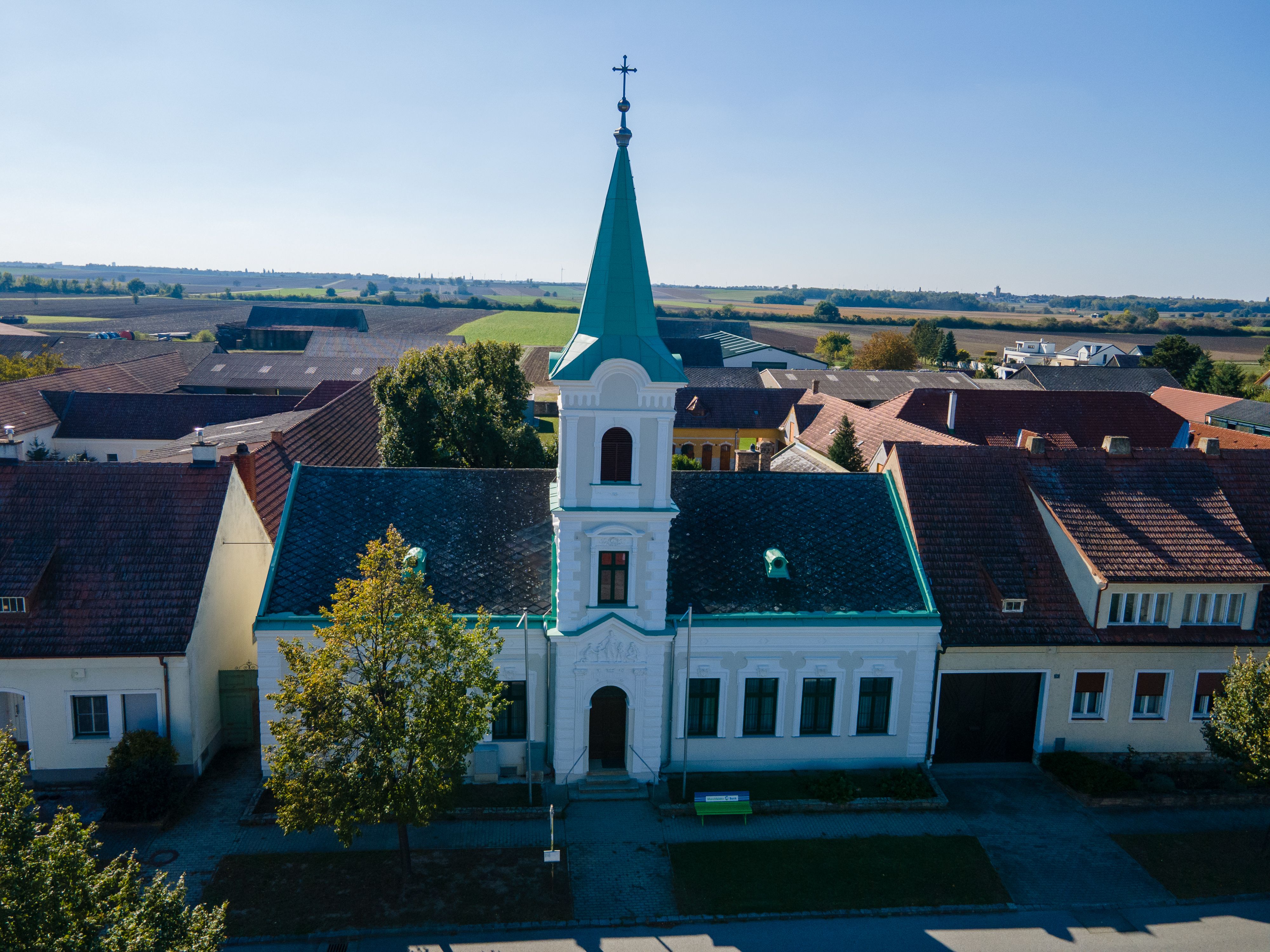 Kostel v obci Weiden an der March se zelenou věží a křížem, obklopený domy a poli.