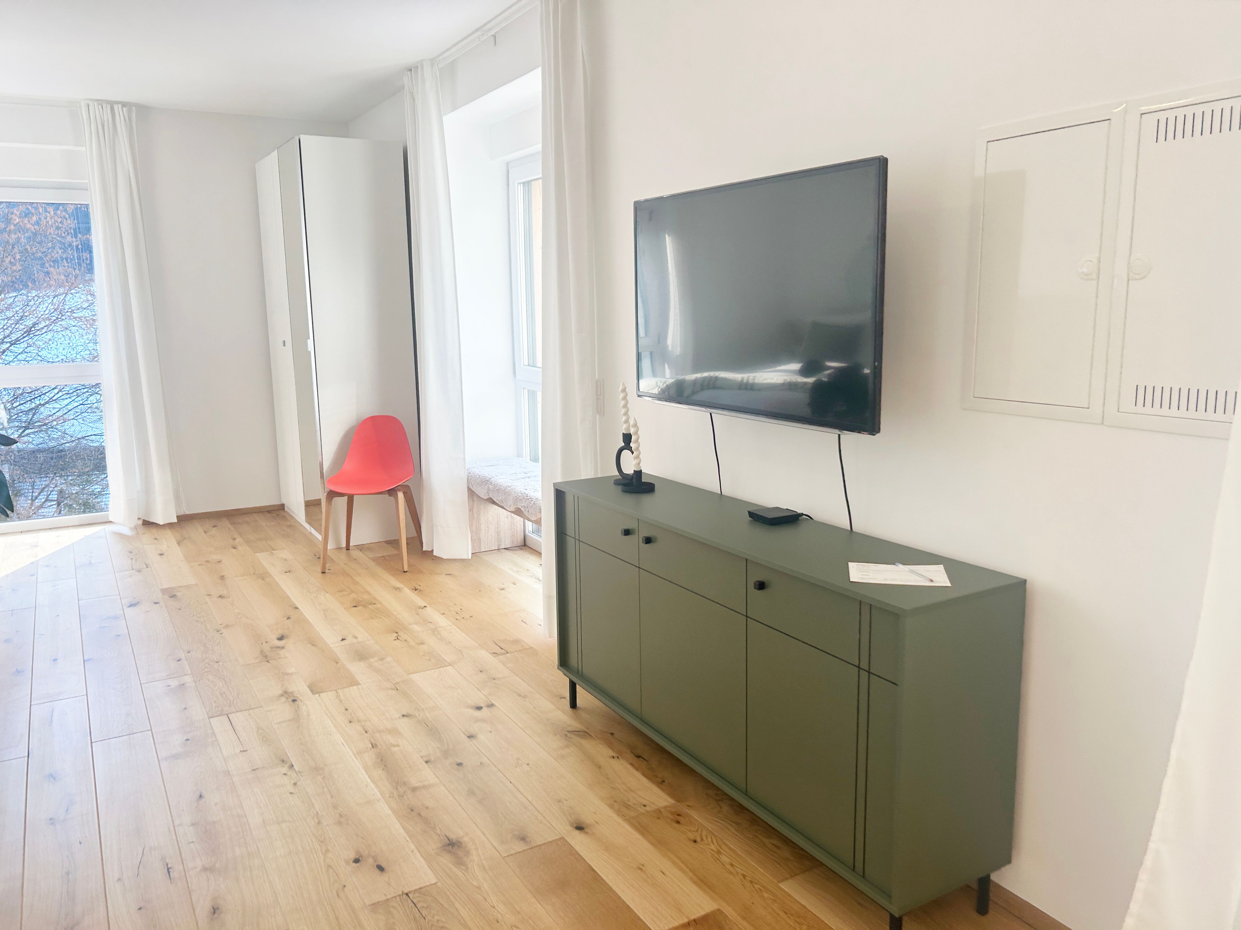 Moderní obývací pokoj s dřevěnou podlahou, zeleným příborníkem, nástěnnou televizí a červeným křeslem před zrcadlem.