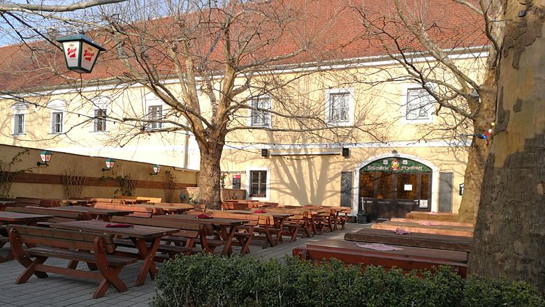 Pivní zahrádka s dřevěnými lavicemi a stoly před žlutou budovou.