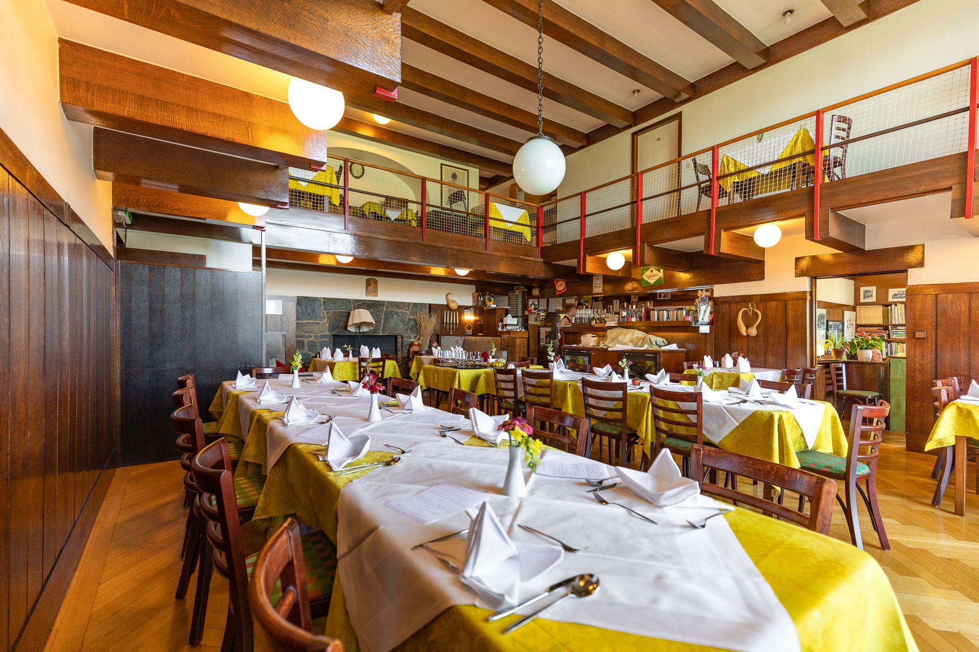 Pohled do interiéru útulné restaurace s dřevěnými trámy, krytými stoly a galerií.