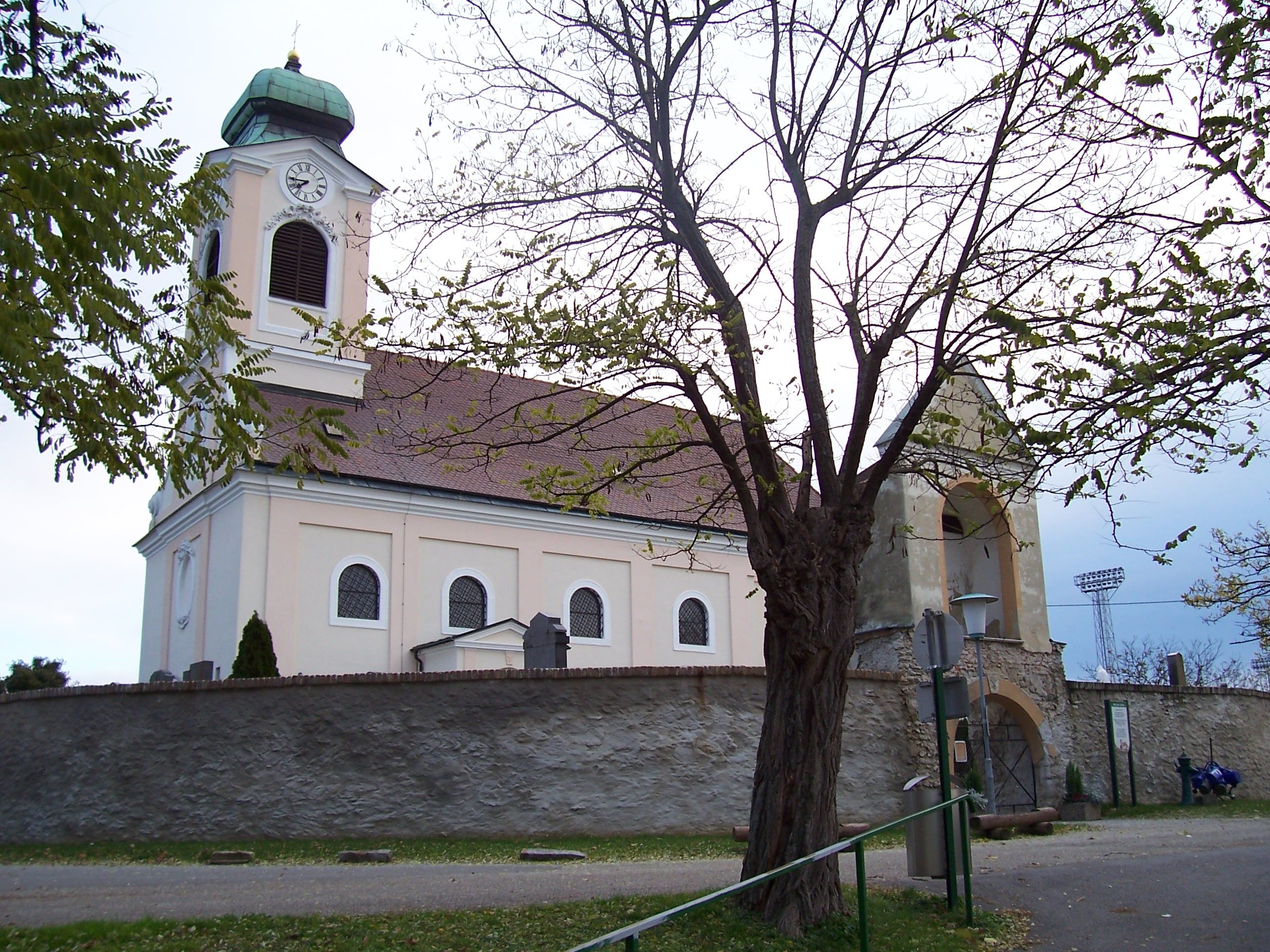 Kostel s věží a hodinami, obklopený stromy a kamennou zdí.