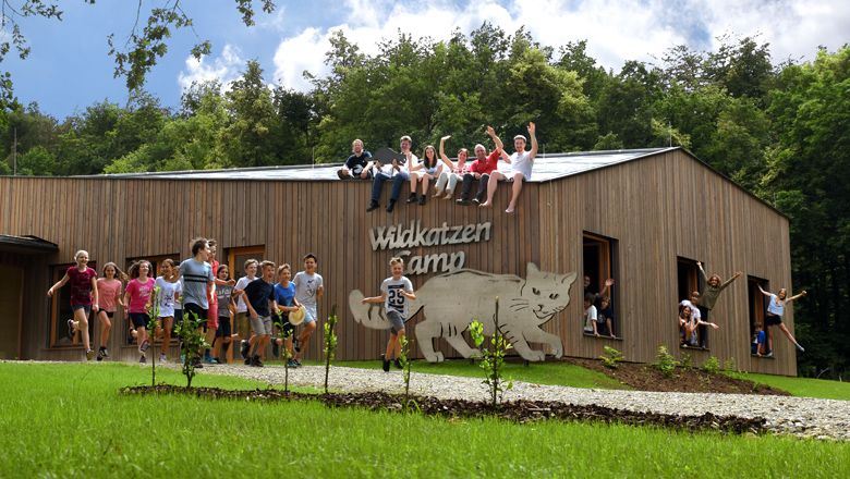 Skupina dětí a dospělých sedí na dřevěné budově s nápisem "Wildcat Camp". Některé děti před ní stojí a skáčou.