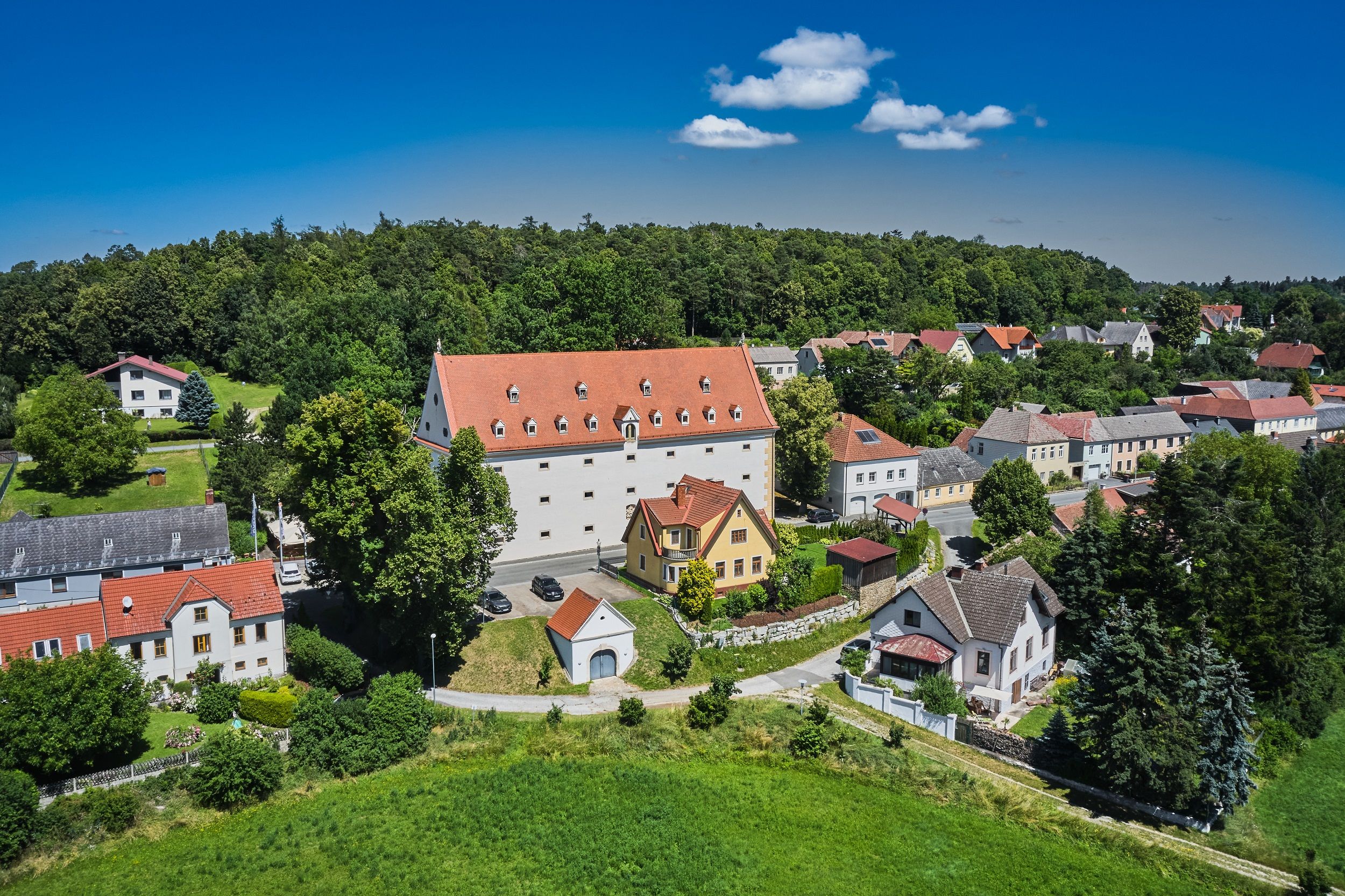 Letecký pohled na vesnici s velkou bílou budovou s červenou střechou, obklopenou menšími domy a spoustou zeleně.