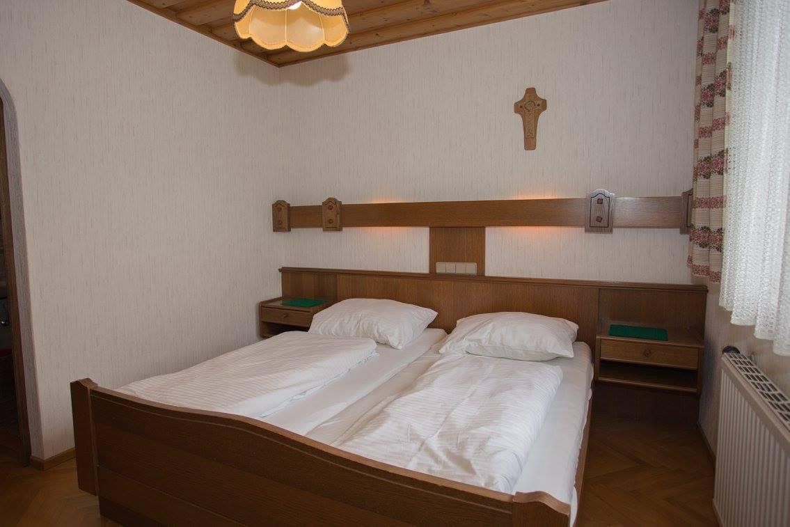 Tradiční dvoulůžkový pokoj s dřevěným nábytkem, dvěma postelemi, křížem na stěně a lampou na stropě.