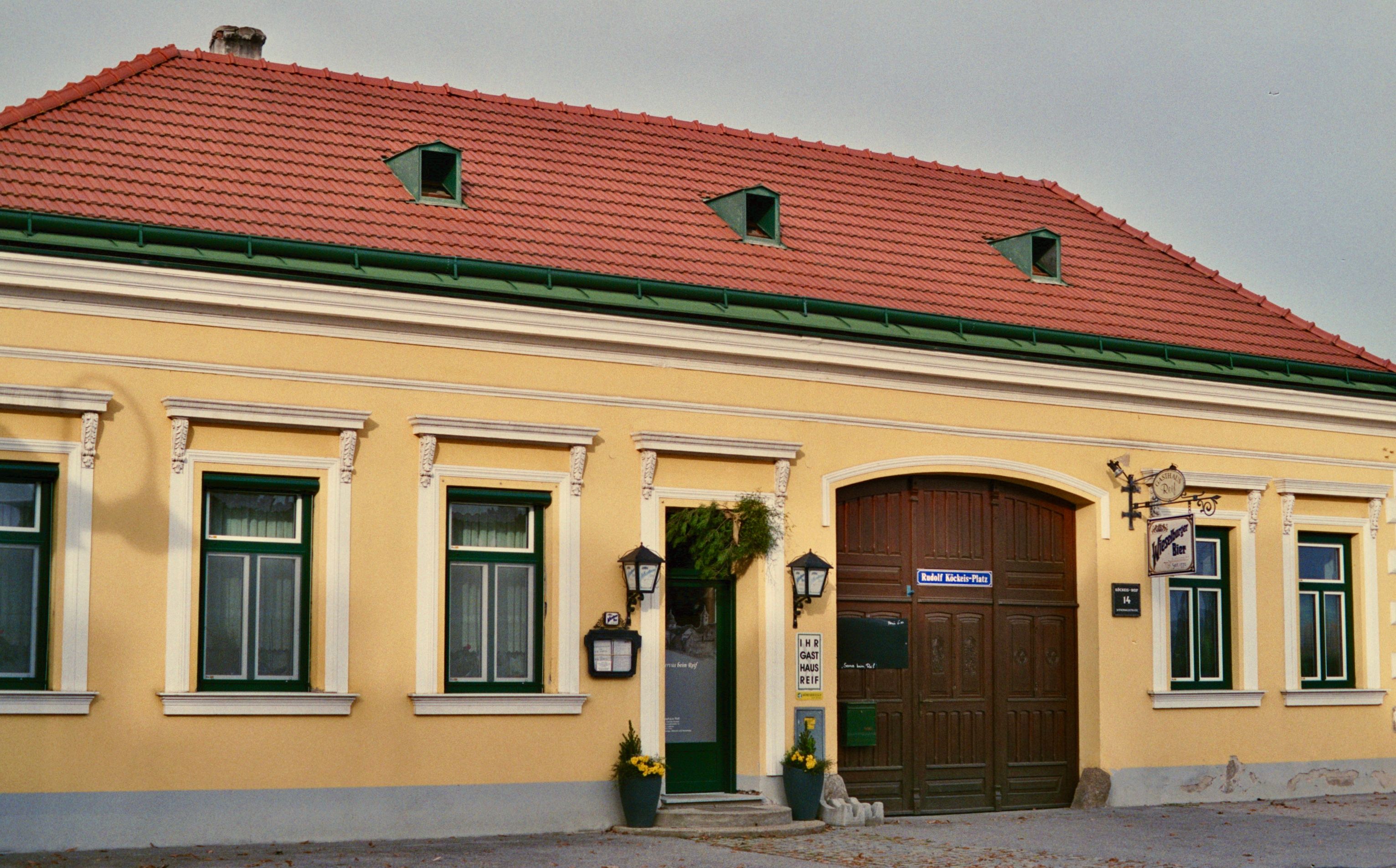 Pohled zvenčí na žlutý hostinec s červenými střešními taškami a zelenými okenními rámy.