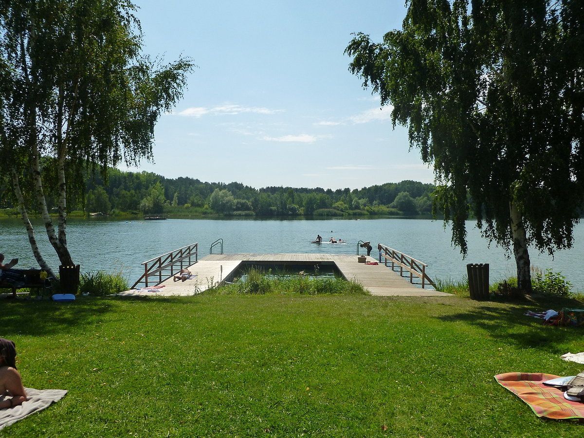 Molo u jezera Bergwerksee Langau s koupajícími se a opalujícími se lidmi. Obklopeno stromy a zelenými loukami.