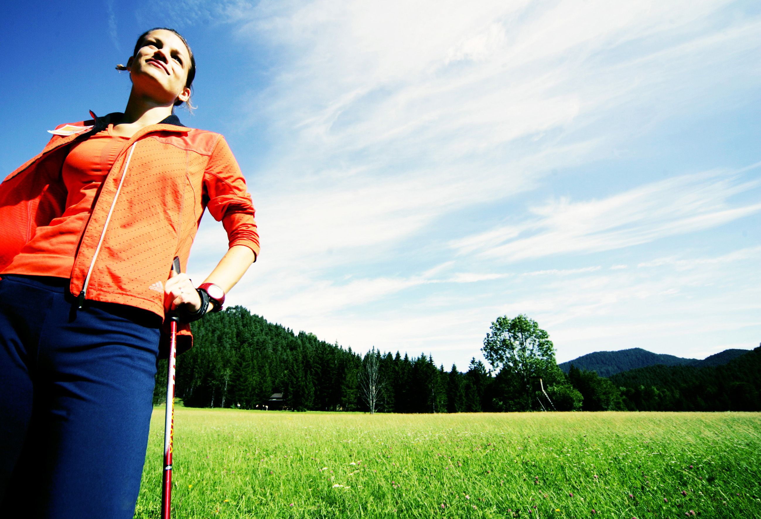 Žena ve sportovním oblečení s holemi na nordic walking na louce před lesem.