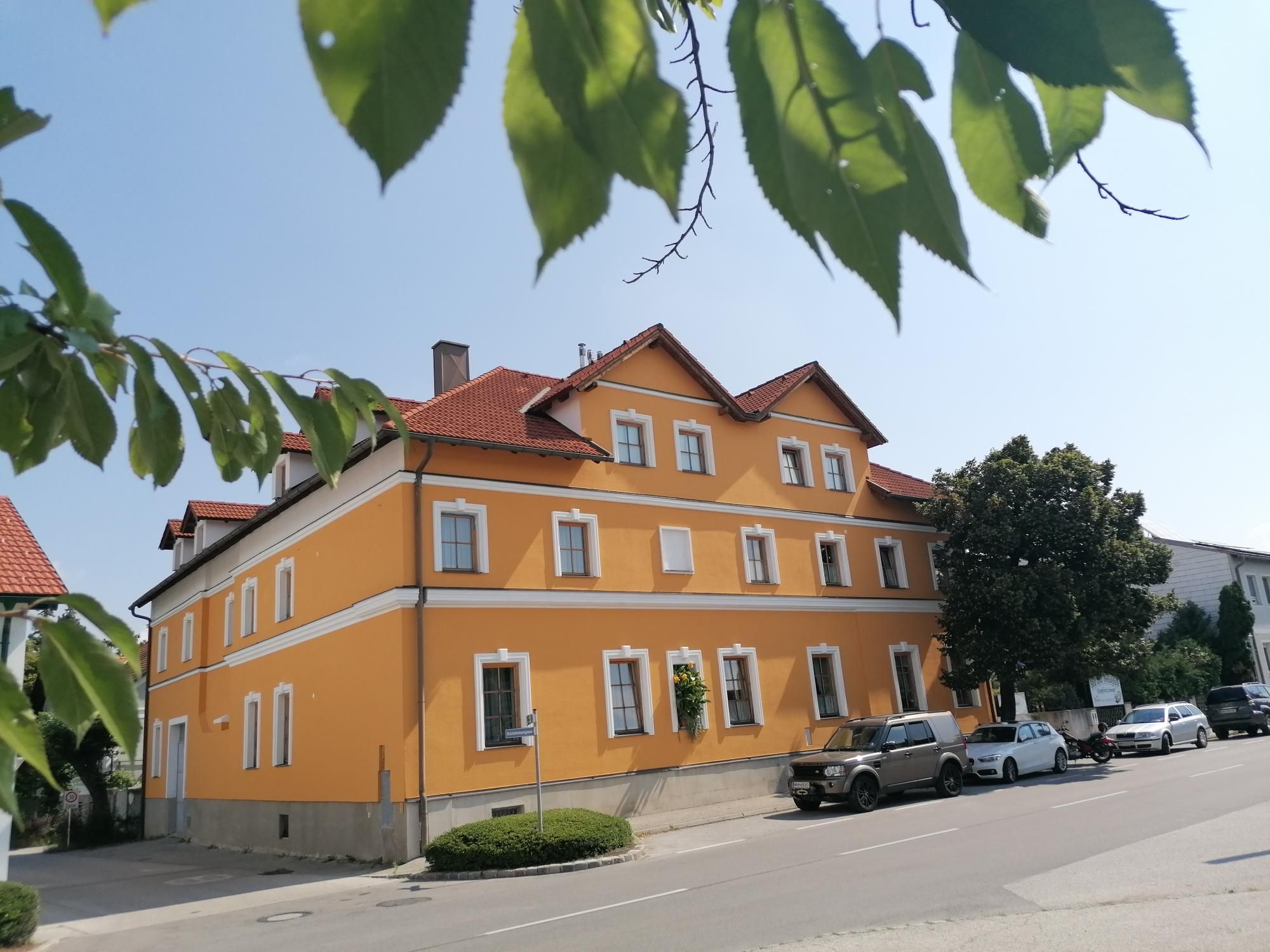 Žlutá budova s červenými střechami, obklopená stromy a zaparkovanými auty.
