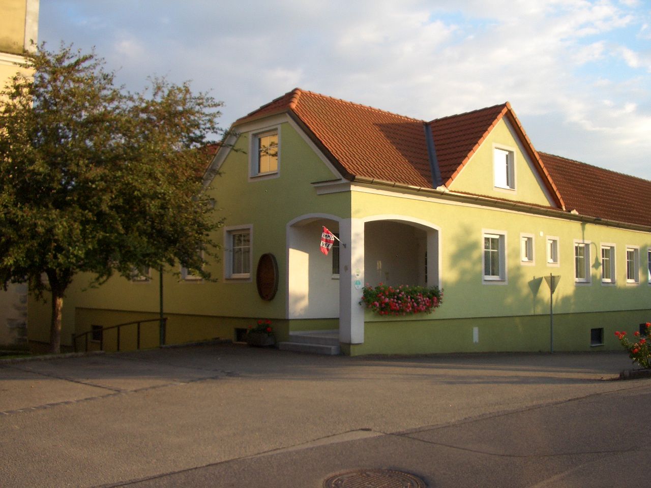 Žlutá budova s červenou střechou a květinami u vchodu.