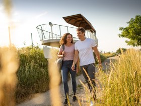 P&aacute;r kr&aacute;č&iacute; po cestě vedle vyhl&iacute;dkov&eacute; plo&scaron;iny na Wagramu, obklopen vysokou tr&aacute;vou a pod jasnou oblohou.