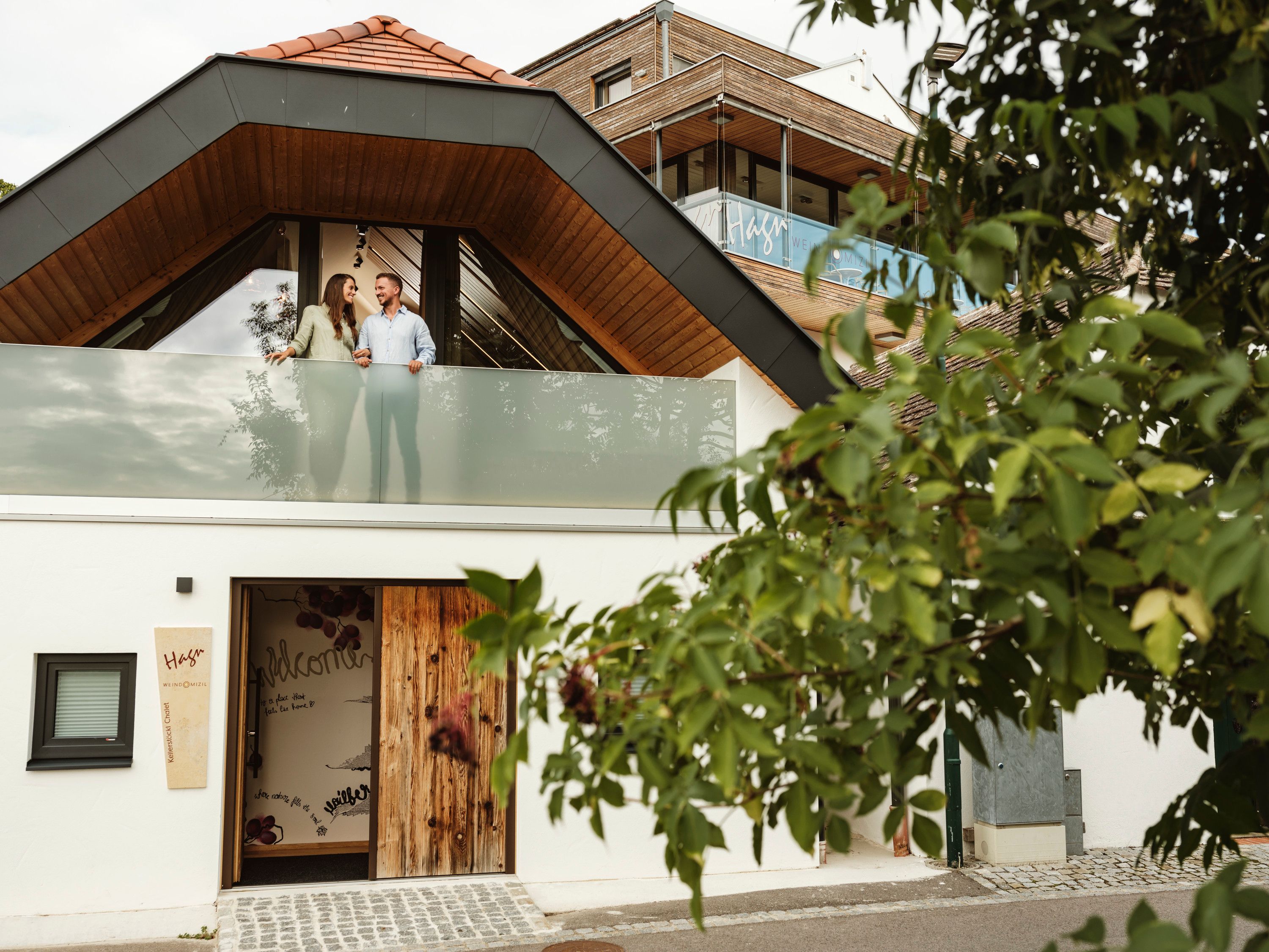 Moderní budova s párem na balkoně, obklopená stromy.