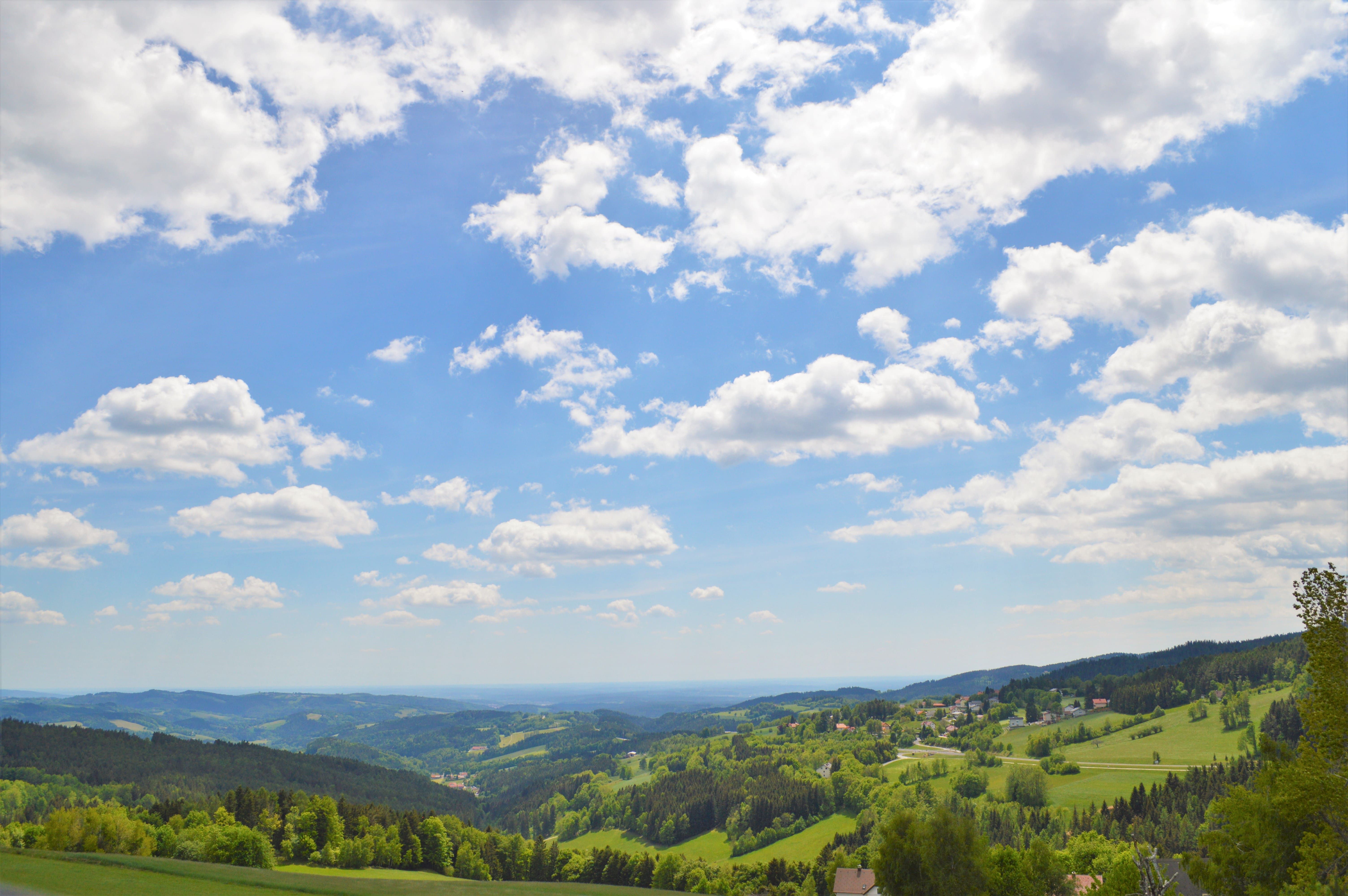 Panoramatický pohled na kopcovitou krajinu s lesy a loukami pod modrou oblohou s bílými mraky.