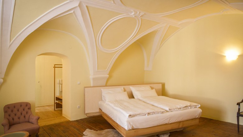 Elegantní ložnice s klenutým stropem, manželskou postelí, křeslem a dřevěnou podlahou.