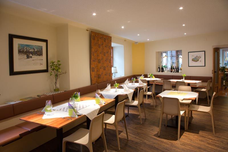 Moderní jídelna s prostřenými stoly, židlemi a dekoracemi. Na stěnách visí obrazy a na okenním parapetu stojí láhve s vínem.