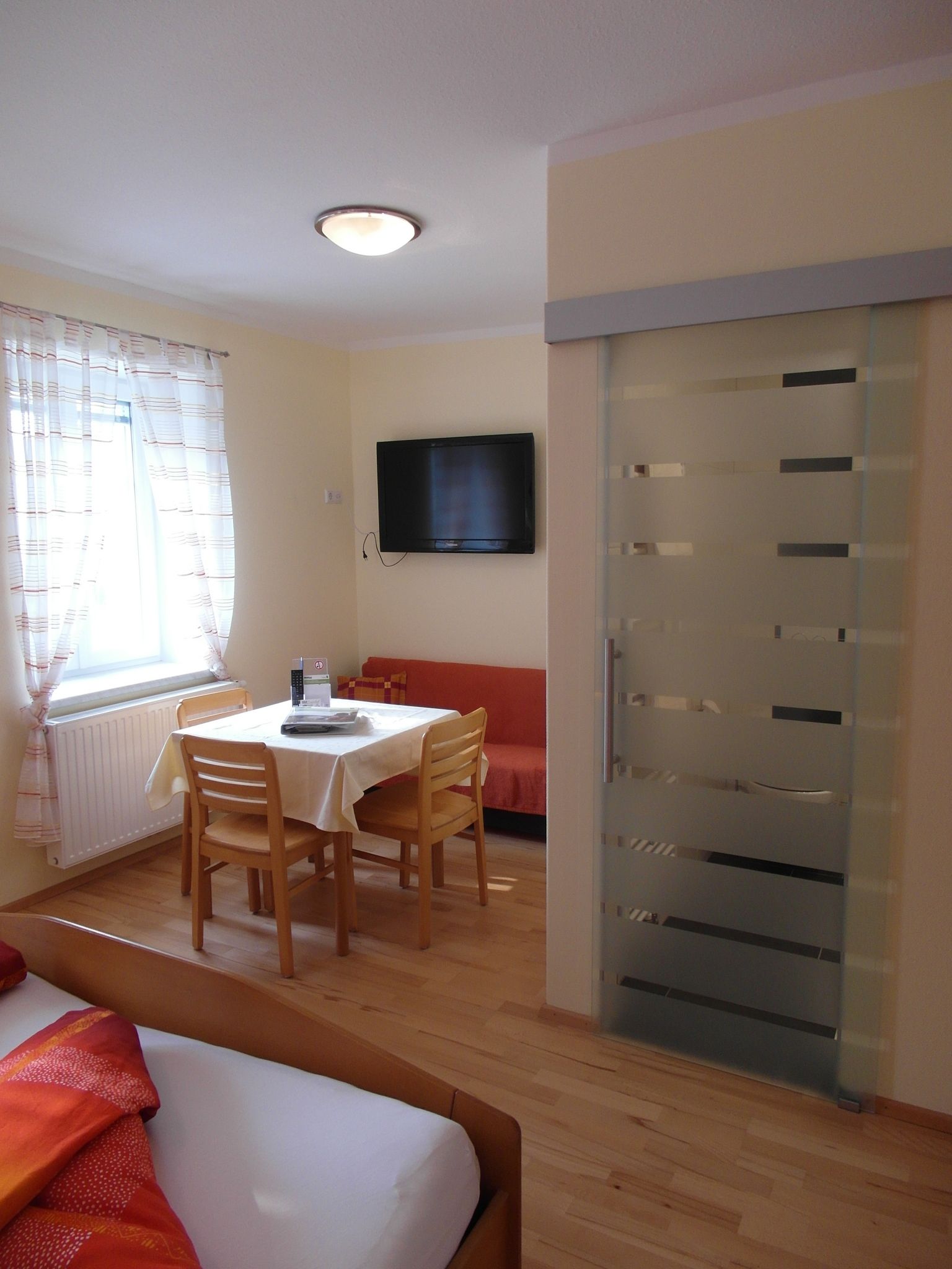 Světlý pokoj s jídelním koutem, stolem, židlemi, postelí a TV s plochou obrazovkou.