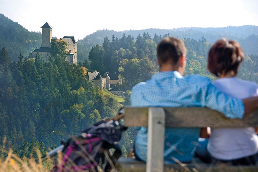 Pár sedí na lavičce s výhledem na hrad v zalesněné krajině.