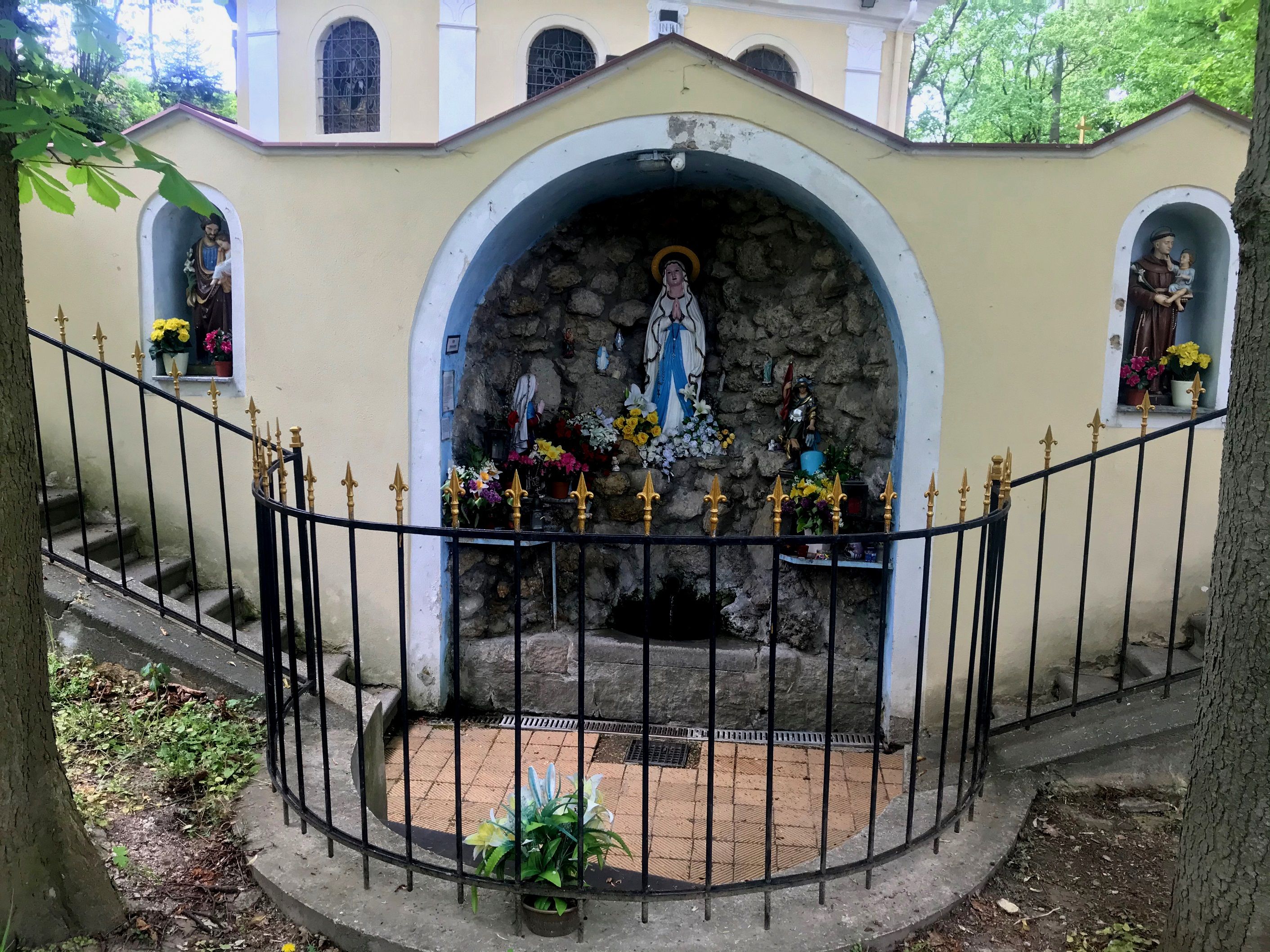 Grotta s náboženskými sochami a květinami, obehnaná kovaným plotem.