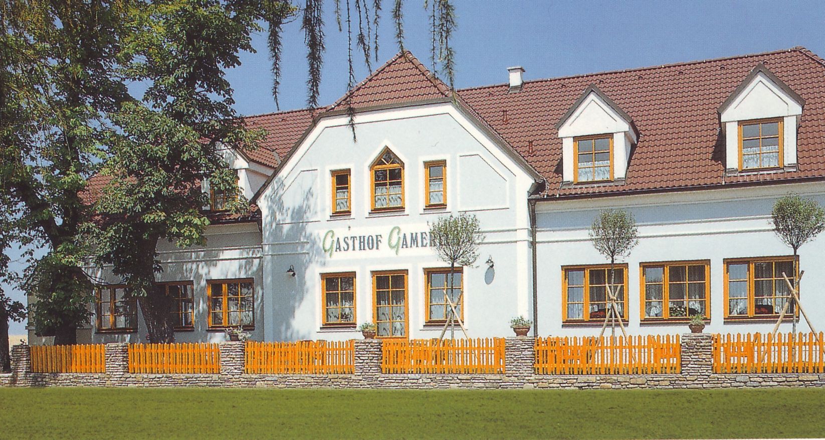 Tradiční hostinec s bílou fasádou a červenými střešními taškami, obklopený stromy a dřevěným plotem.