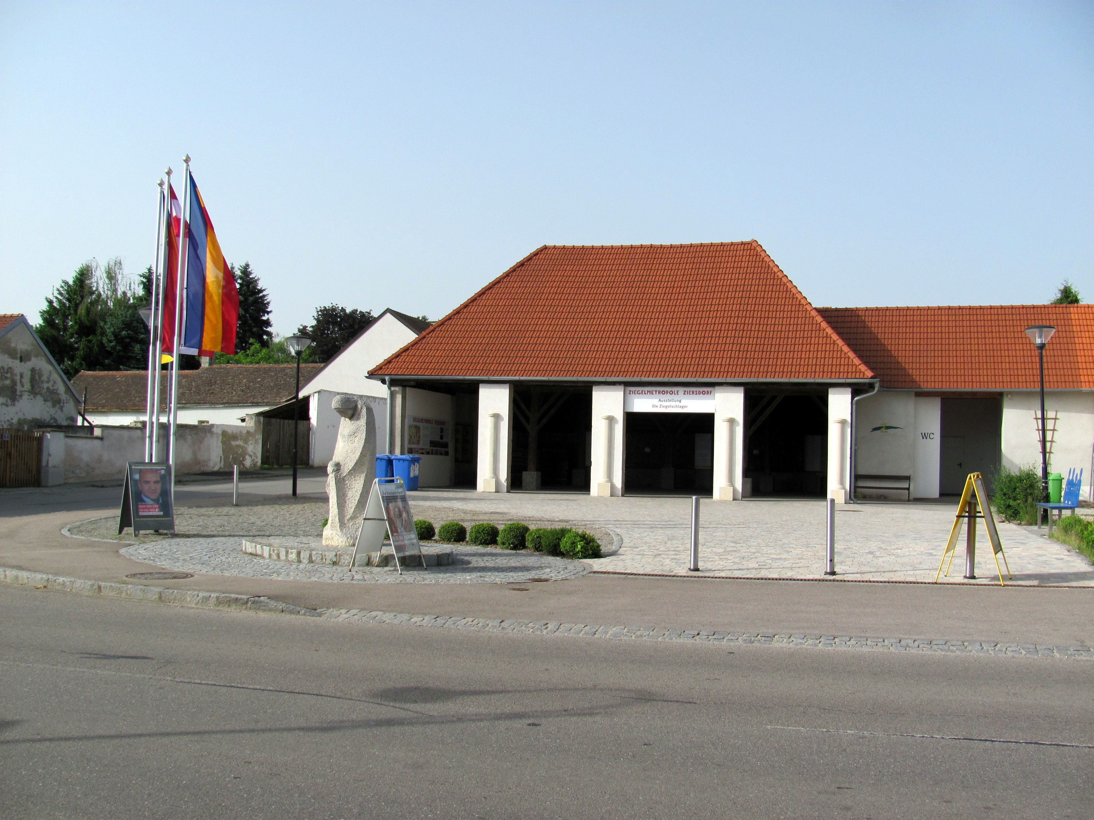 Pohled zvenčí na cihlové muzeum v Ziersdorfu s červenými střešními taškami a vlajkami.