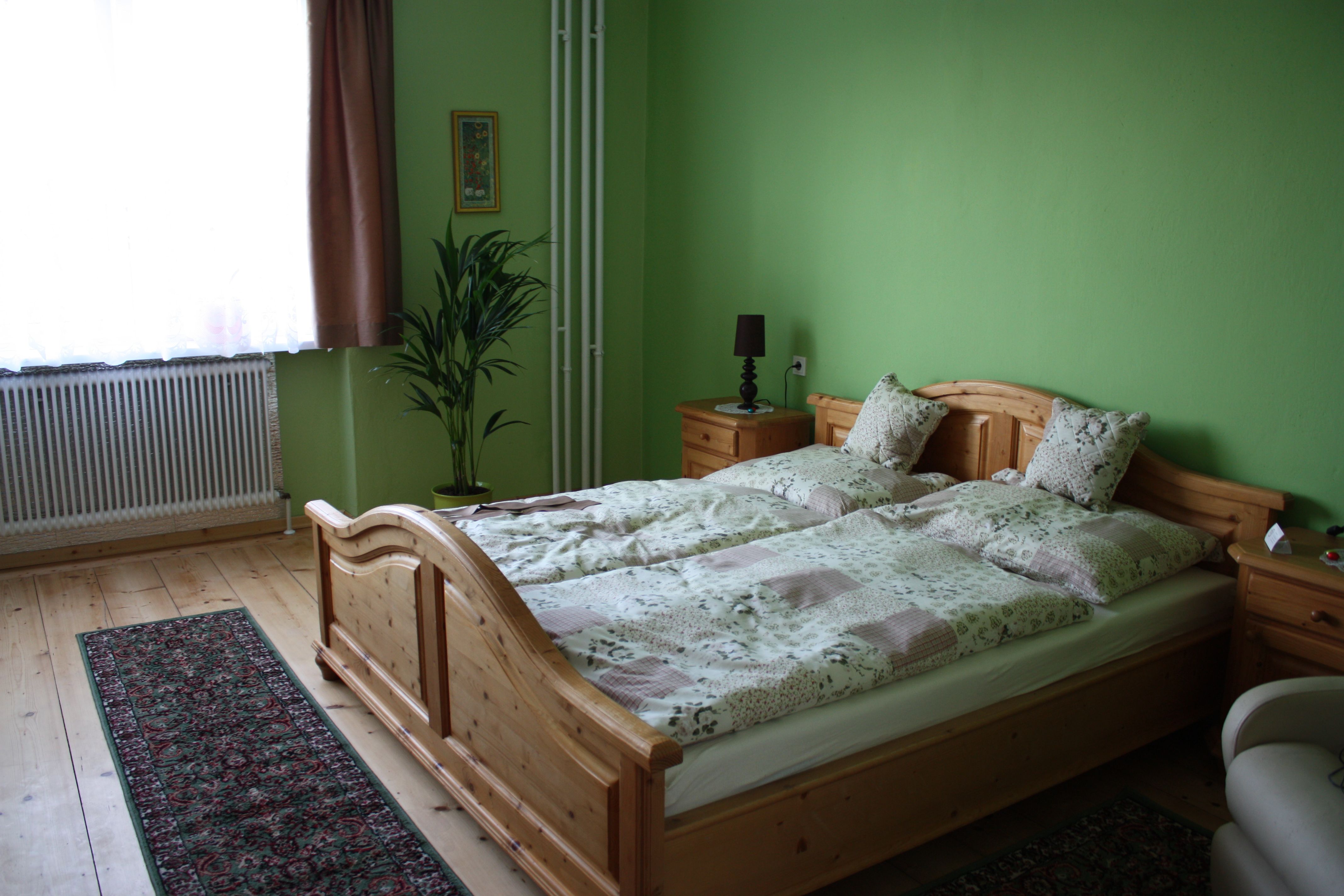 Útulný pokoj pro hosty se zeleně natřenými stěnami, dřevěnou postelí, nočními stolky, rostlinou a kobercem na dřevěné podlaze.