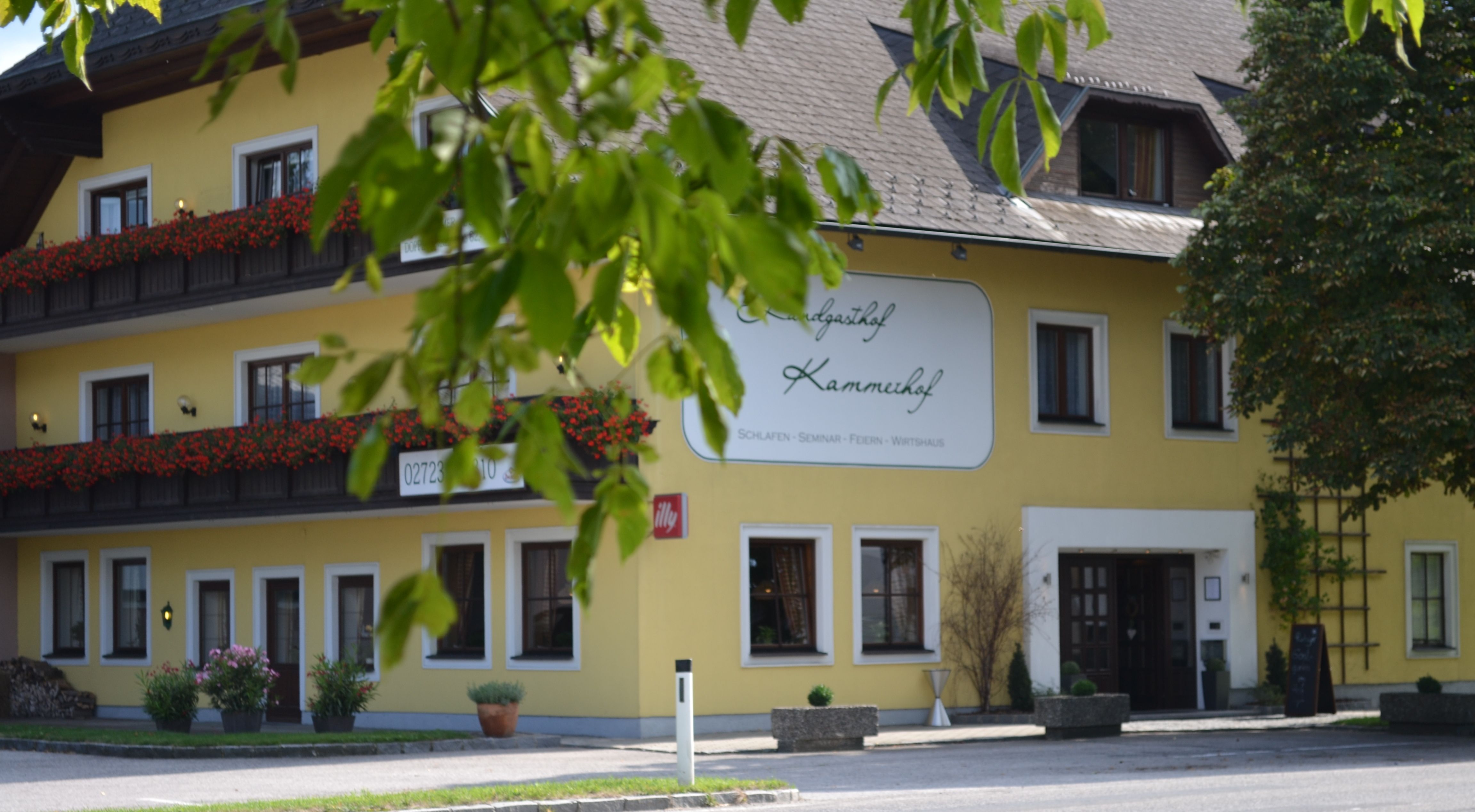 Žlutá budova s nápisem "Landgasthof Kammerhof", květinové truhlíky a stromy v popředí.