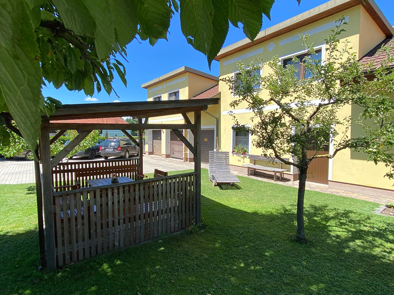 Zahrada s pavilonem, žlutým domem a autem v pozadí.