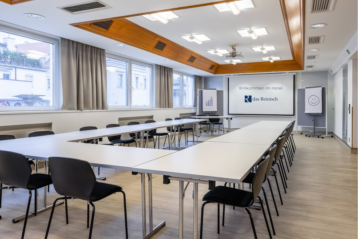 Seminární místnost v hotelu Das Reinisch se stoly, židlemi a prezentačním plátnem.