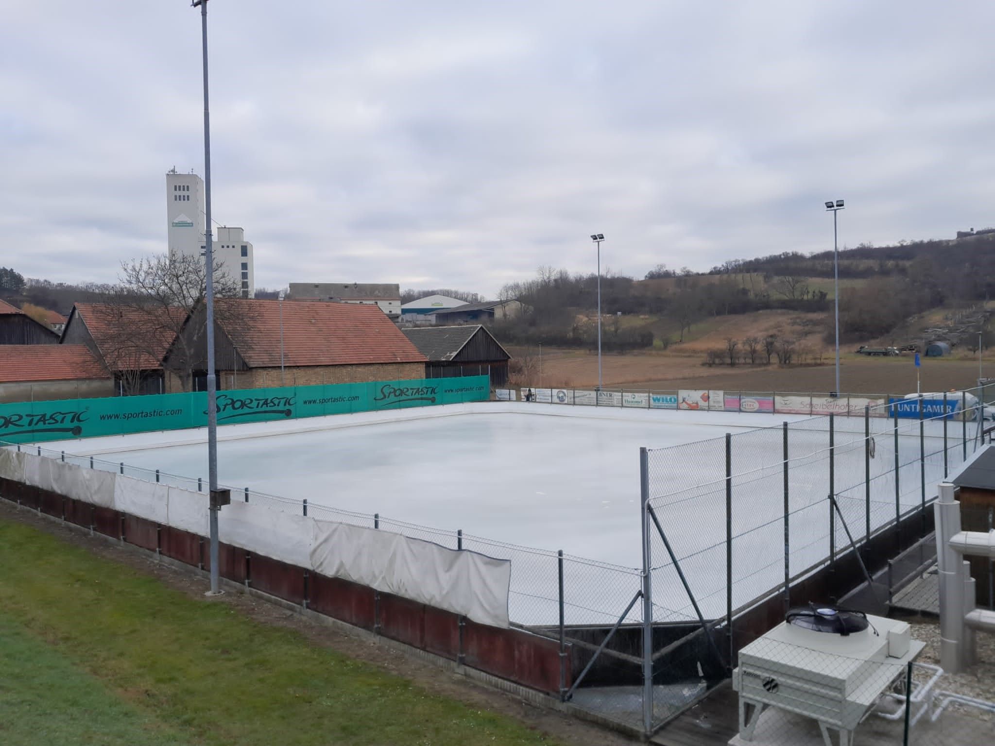 Prázdné kluziště s okolními budovami a kopci v pozadí.