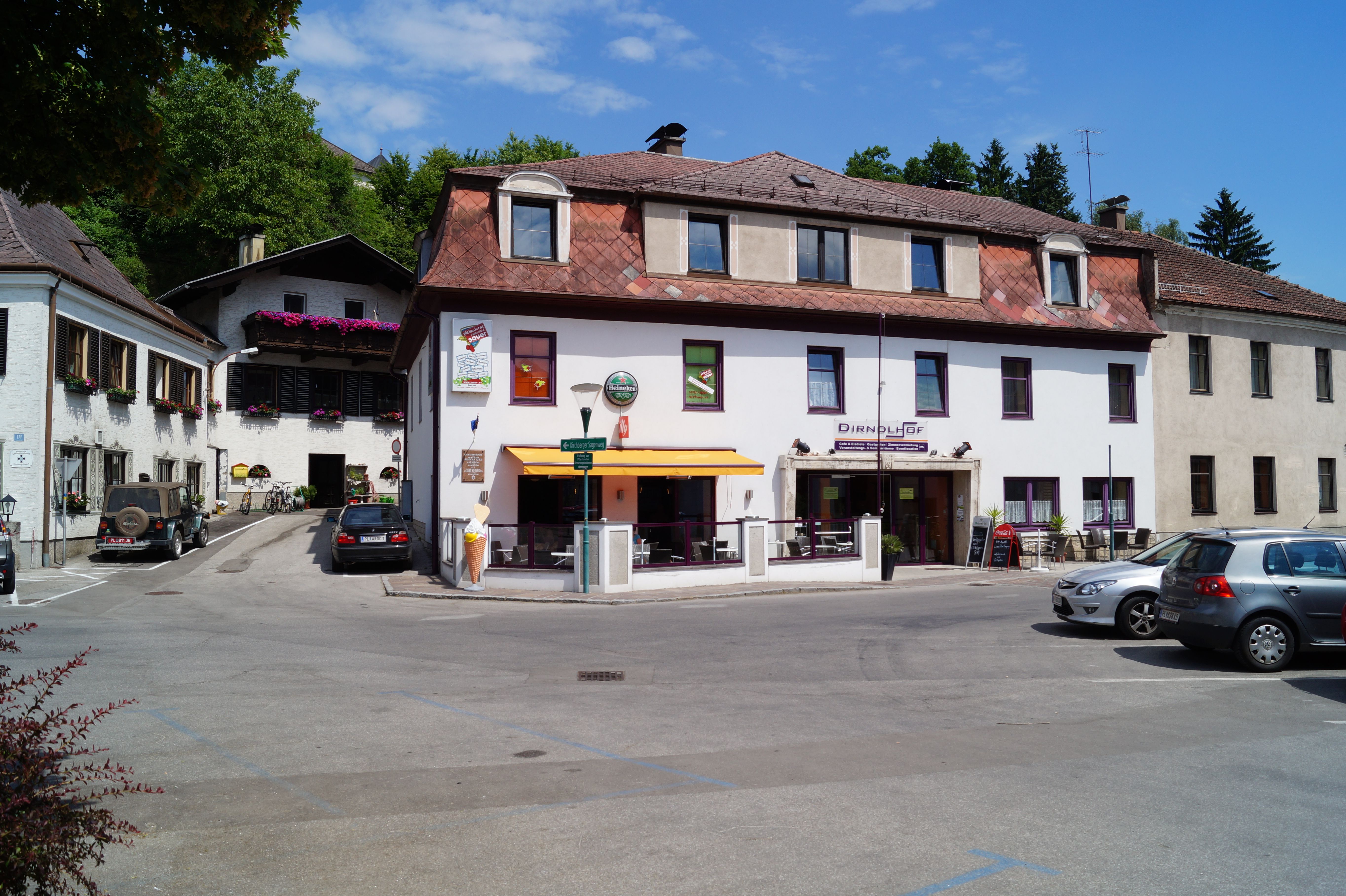 Budova s nápisem "Dirndlhof" a terasa se slunečníkem. Auta parkují na parkovišti před ní.