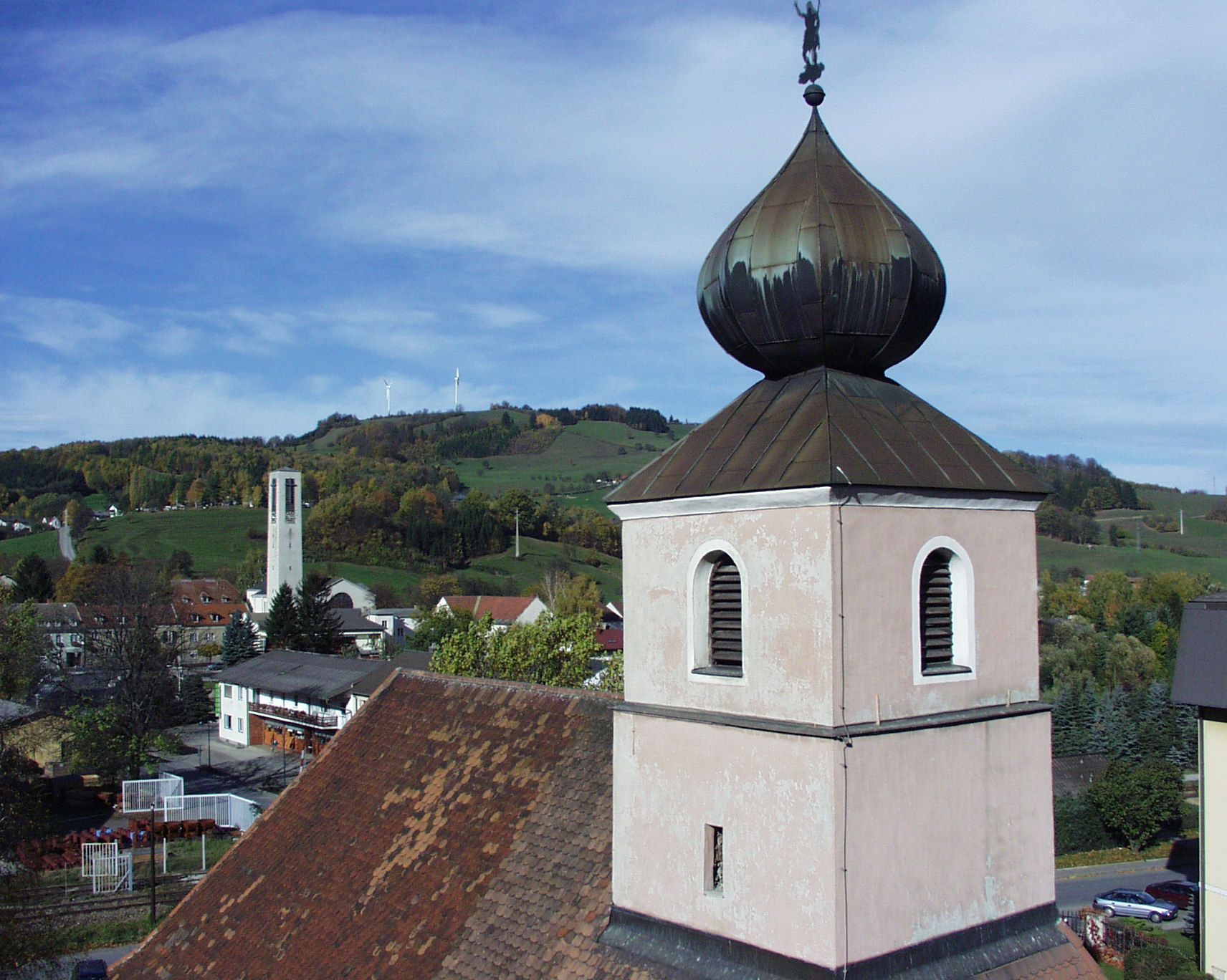 Kostelní věž s cibulovou střechou před kopcovitou krajinou a větrnými turbínami.