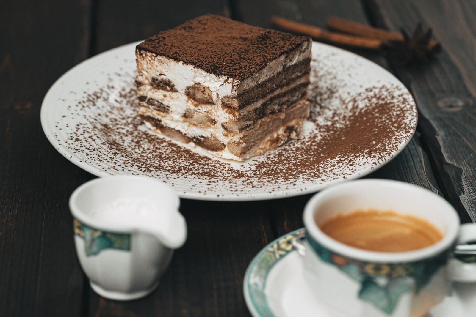 Na dřevěném stole stojí talíř s kouskem tiramisu, vedle něj šálek kávy a konvička s mlékem.