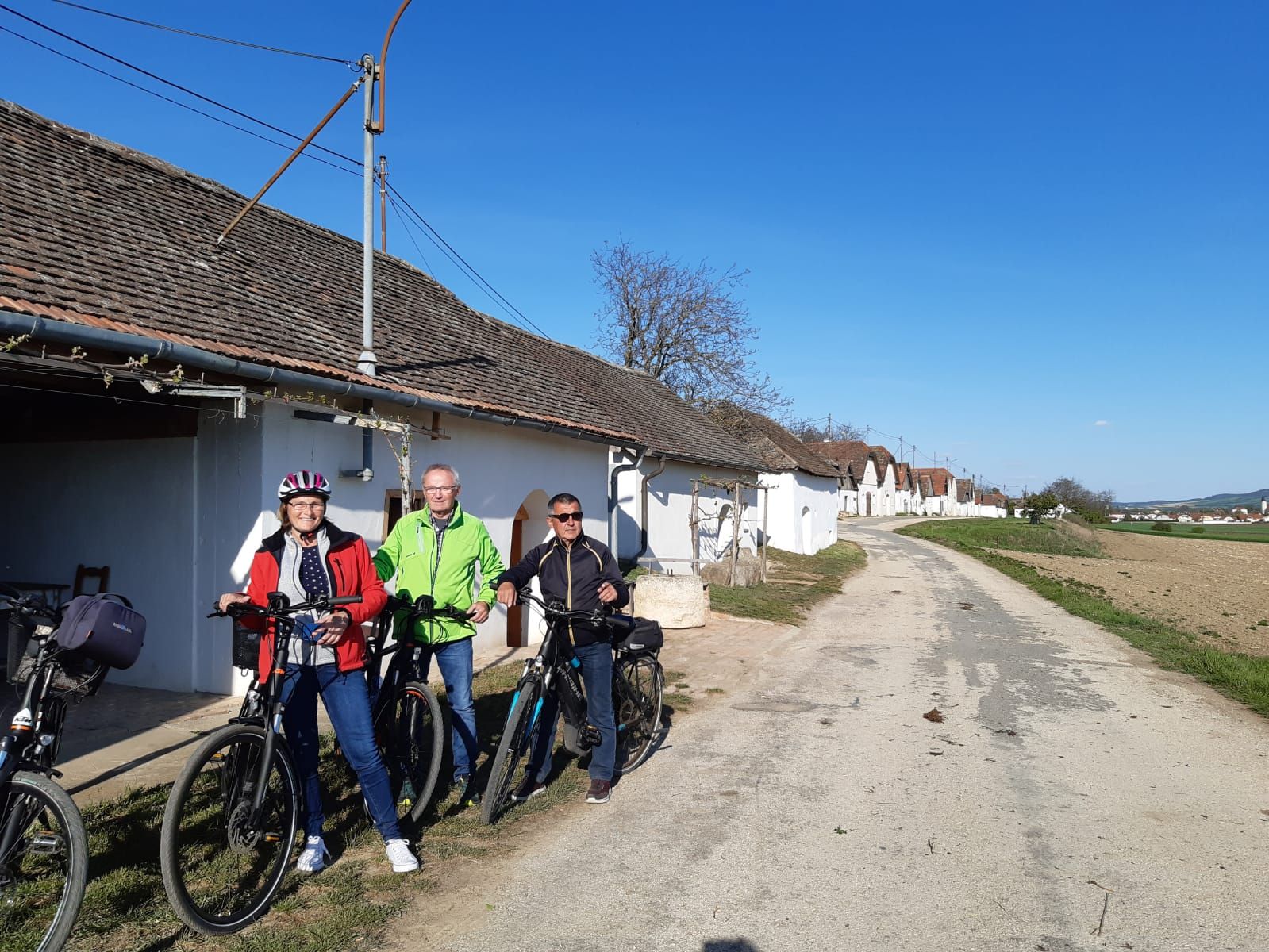 Tři lidé s koly před řadou vinných sklepů na venkově.