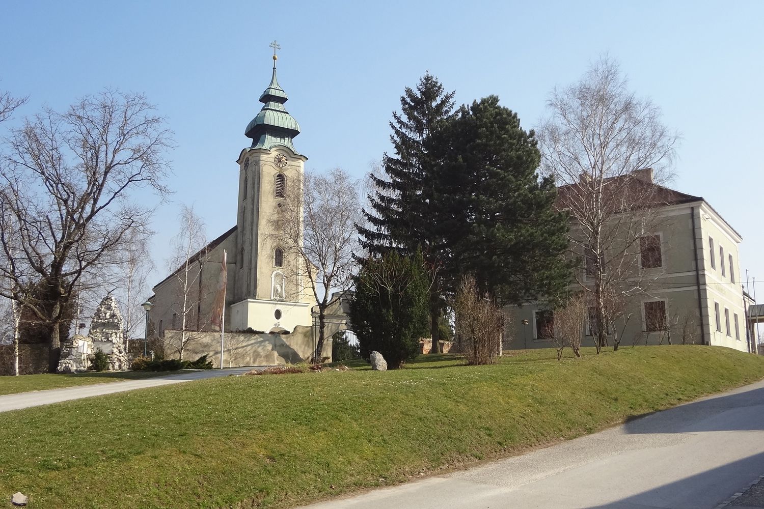 Kostel s věží a sousední budovou v Gnadendorfu, obklopený stromy a trávníkem.