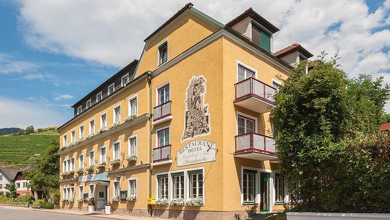 Žlutá hotelová budova s balkony a rostlinami, modrá obloha v pozadí.