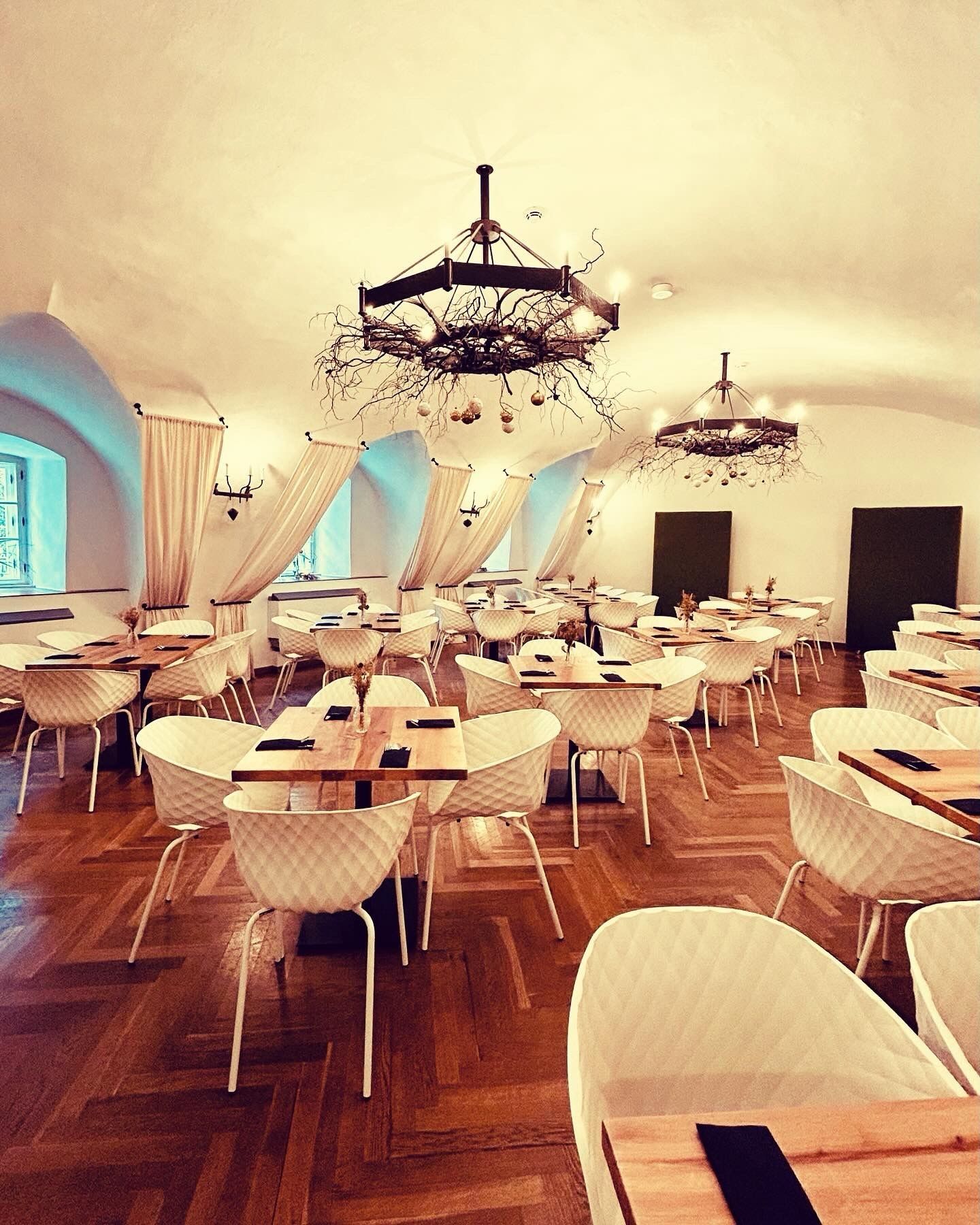 Elegantní jídelna s parketovou podlahou, bílými židlemi, dřevěnými stoly a ozdobnými lustry.