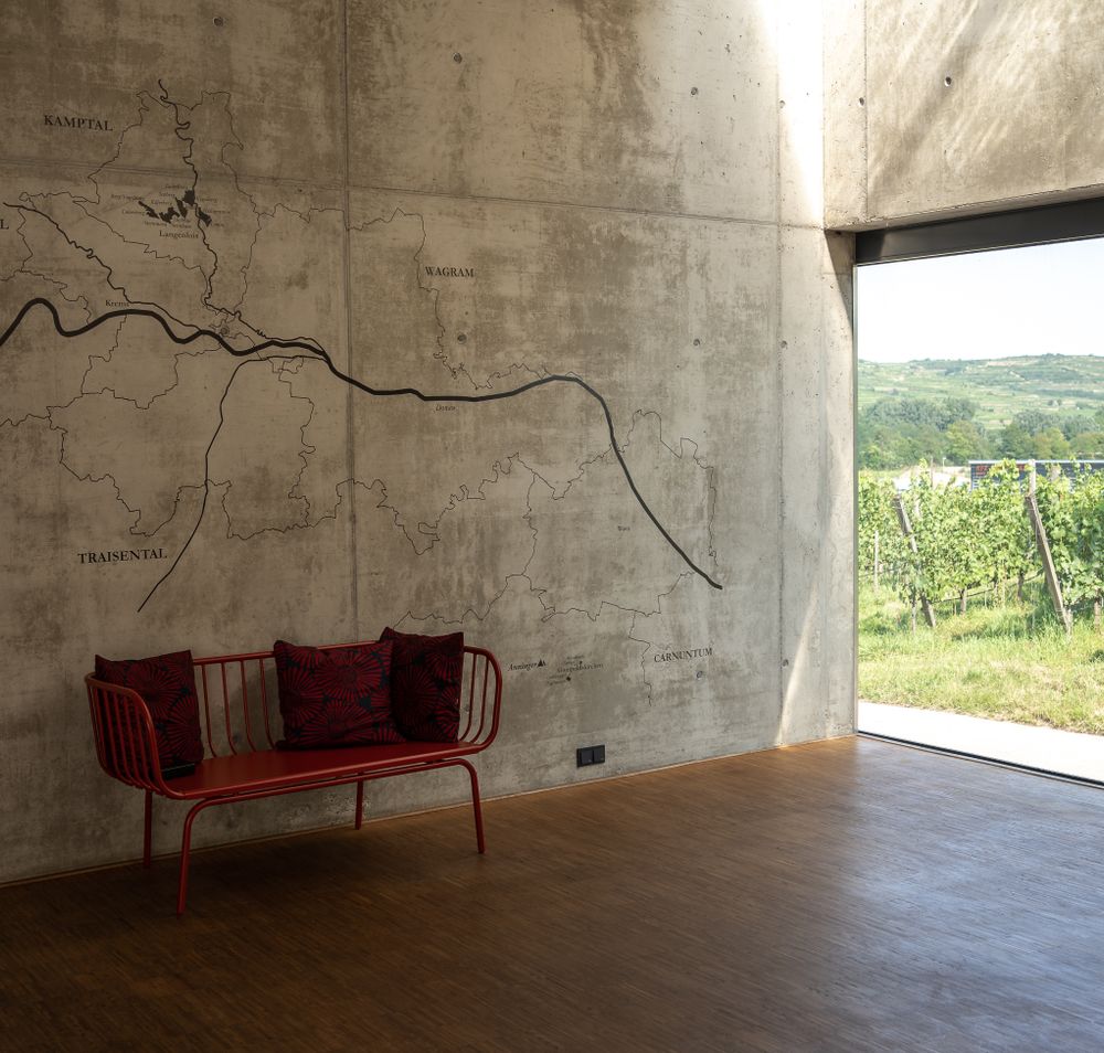 Ein einladender Raum mit einer stilvollen roten Bank und einer beeindruckenden Wandkarte, die die Weinregion zeigt. Durch das große Fenster strömt das Licht herein und bietet einen malerischen Blick auf die sanften Hügel und Weinreben, die in der warmen Sonne leuchten.