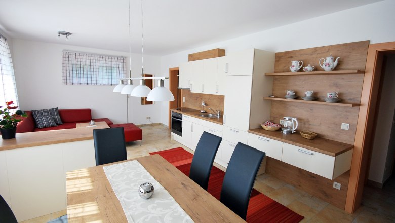 Modern&iacute; ob&yacute;vac&iacute; a j&iacute;deln&iacute; prostor s otevřenou kuchyn&iacute;, červenou pohovkou, dřevěn&yacute;m stolem a b&iacute;l&yacute;mi z&aacute;věsn&yacute;mi lampami.