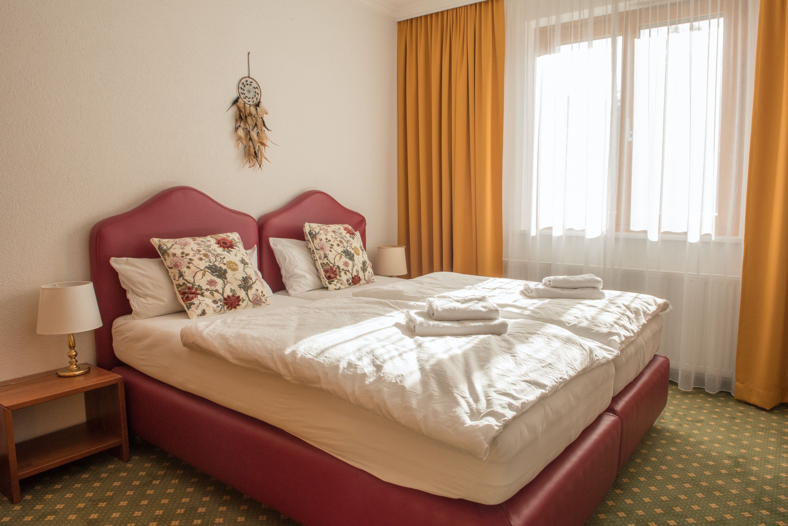 Útulný dvoulůžkový pokoj se dvěma postelemi, žlutými závěsy a lapačem snů na zdi.